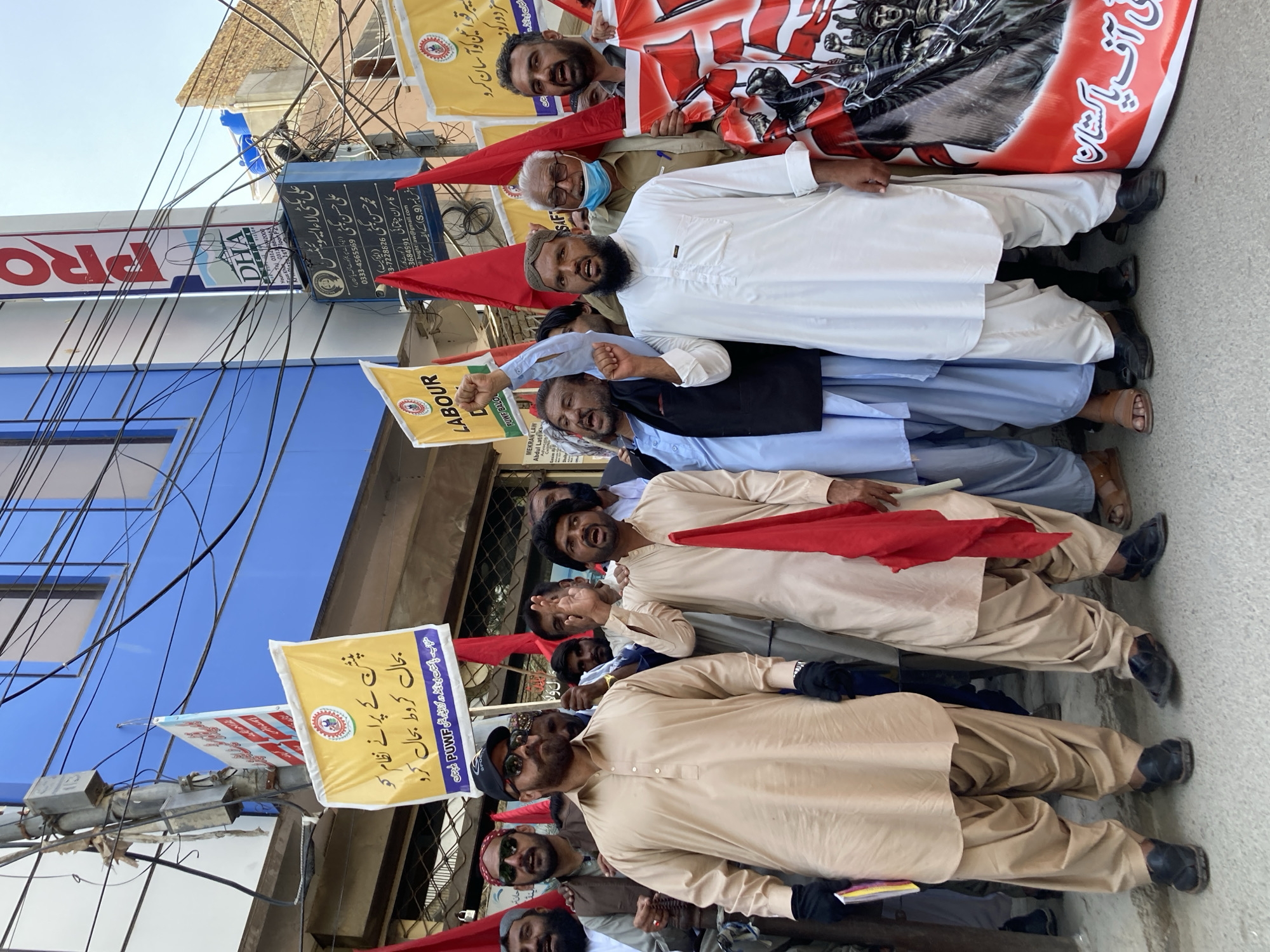A group of men are lined up shoulder-to-shoulder on a street in Quetta City Tehsil, Pakistan. The men are diverse in age, and some are wearing traditional Pakistani clothing, such as shalwar kameez. Several men are holding up red flags and banners with political slogans written in Urdu. Signs for local businesses are visible in the background, including DHA and Ali Bugti Law Associates. Above the shops are a dense web of electrical cables. The men appear to be participating in a labor protest or demonstration.
