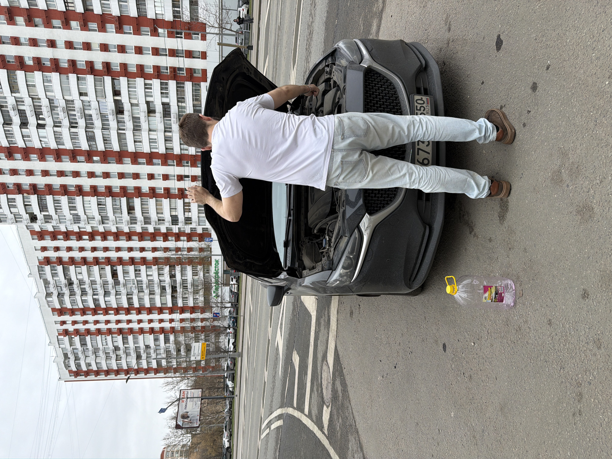A man in a white t-shirt, light-colored jeans, and brown shoes is working on a black car with its hood open. A large plastic bottle with a yellow cap and a pinkish liquid inside is on the ground near the car. The car has a Russian license plate. In the background, there is a tall building with a distinctive white and red striped pattern. The scene is set on a city street in Khimki, Russia.