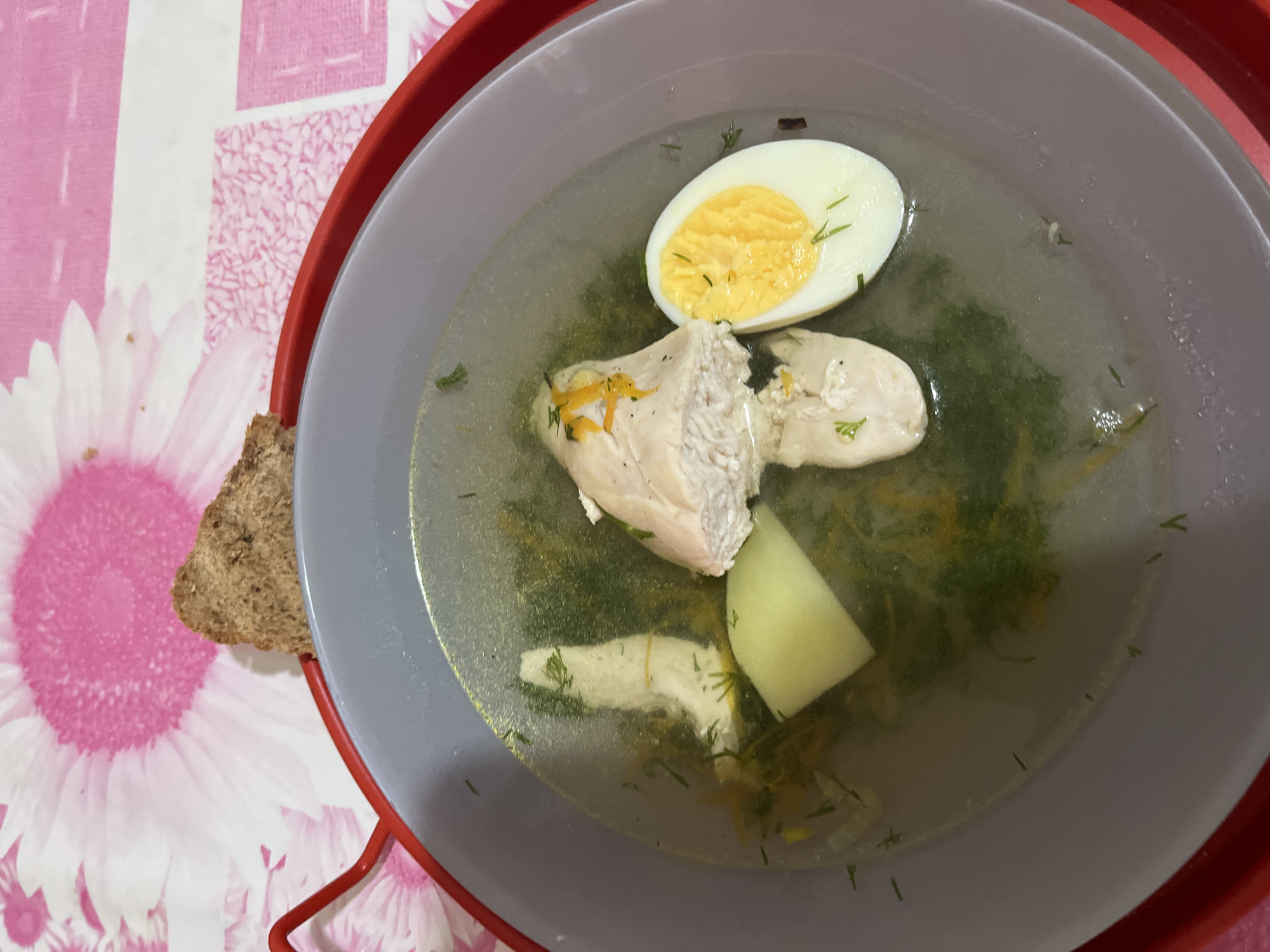 A top-down shot depicts a grey bowl of soup on a red tray, resting on a table with a pink floral pattern. The soup contains a halved hard-boiled egg, pieces of chicken, a chunk of potato, and what appears to be dill or other green herbs. A piece of dark bread sits next to the bowl. The scene doesn't offer clues to confirm it's located in Alanya, Turkey. No people are visible.