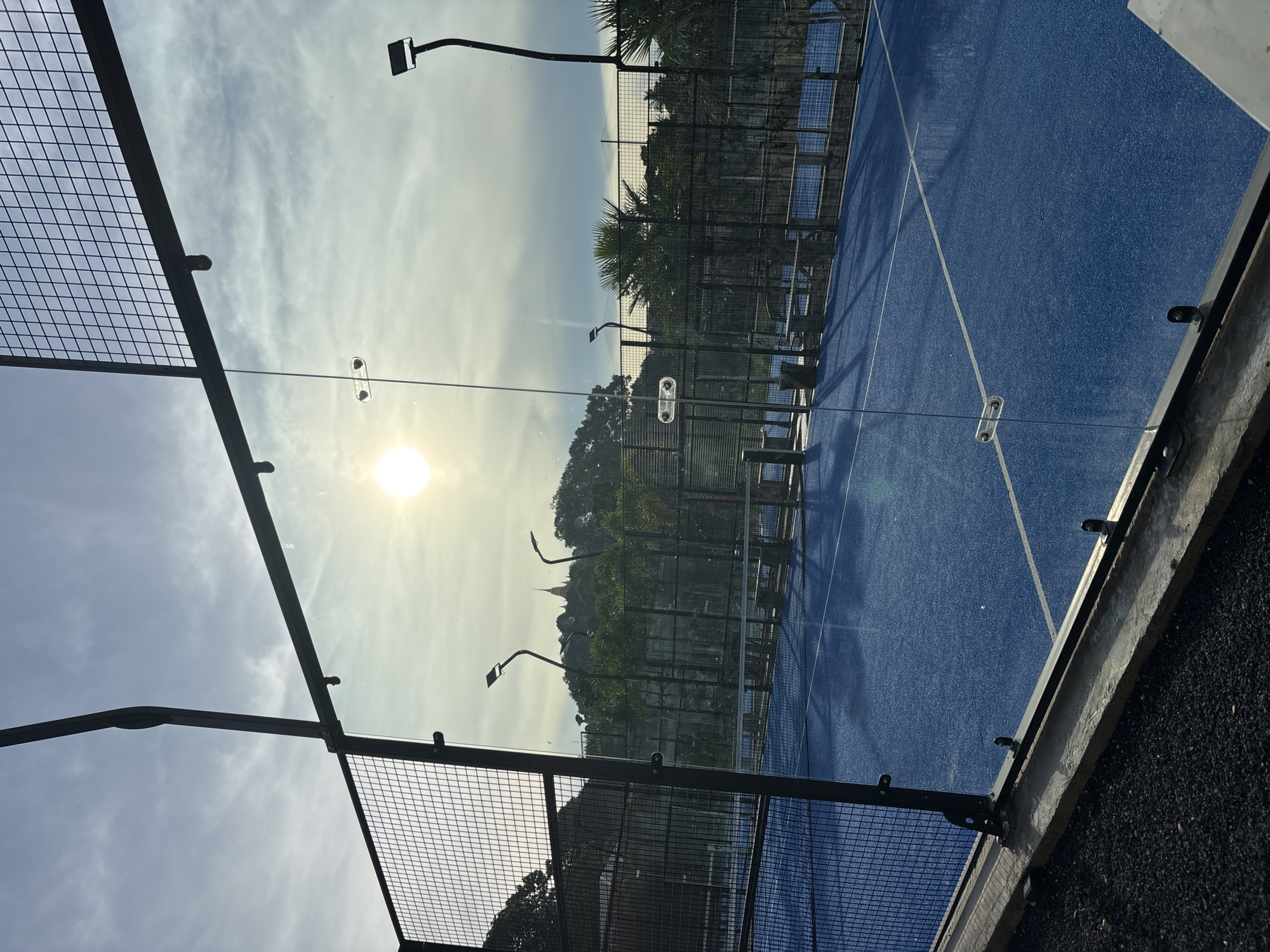 The image shows a blue paddle tennis court in บ้านบางรักษ์, Thailand, with a view through a glass wall and wire fence. The sun is visible in the sky, partly obscured by clouds. There are palm trees and a hill in the background, possibly indicating the location is near a coastal area. Street lamps are visible around the court. The image is slightly rotated.