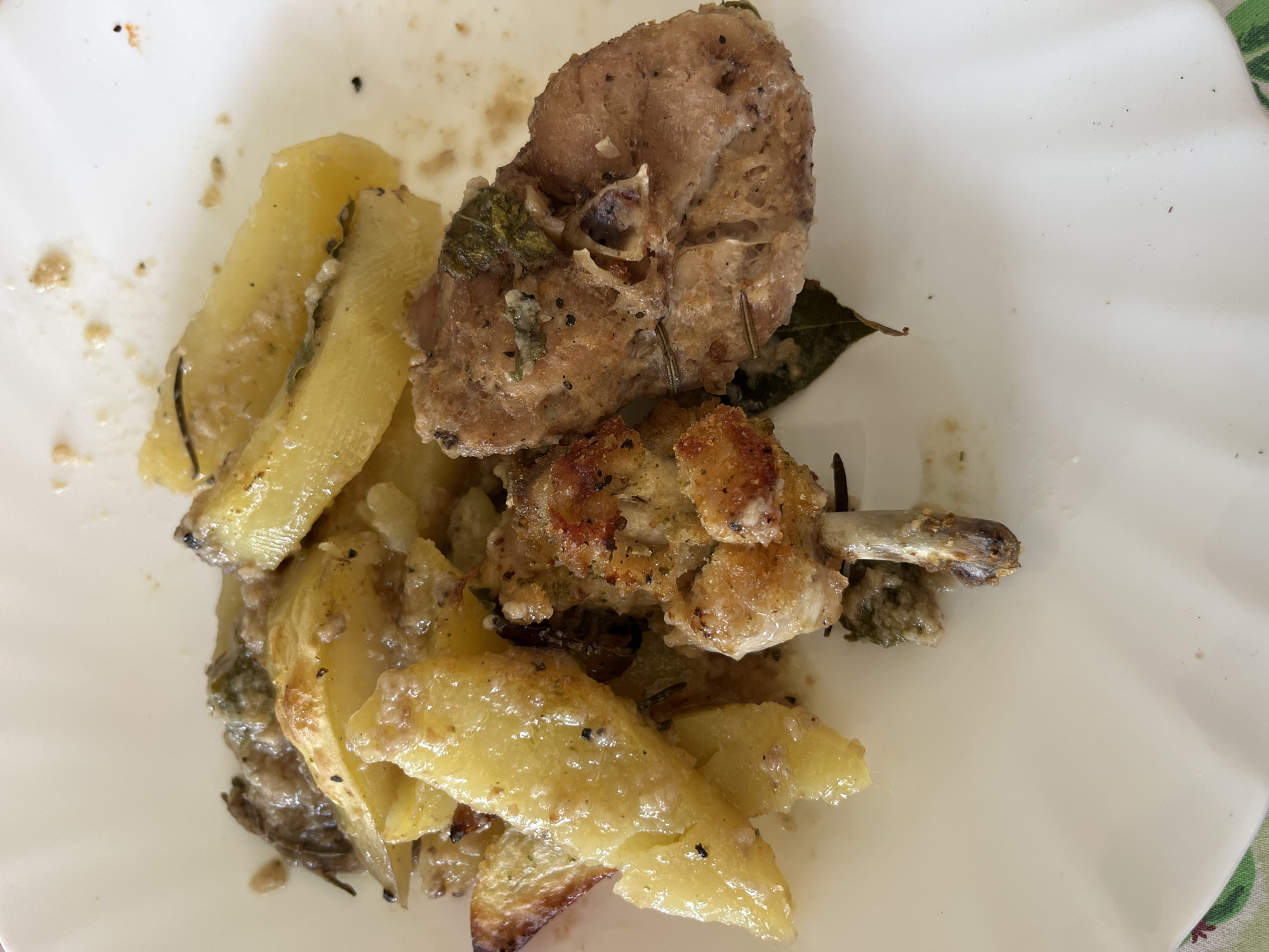 The image shows a plate of food. The food appears to be baked chicken and potatoes. The potatoes are cut into wedges and have a slightly browned and glistening appearance, suggesting they were baked with some kind of fat or oil. The chicken is in two pieces. One piece appears to be a breast or thigh and the other is a drumstick. There are herbs on the chicken and potatoes. The plate is white and appears to be porcelain. Based on information provided, the location is Aradeo, Italy.