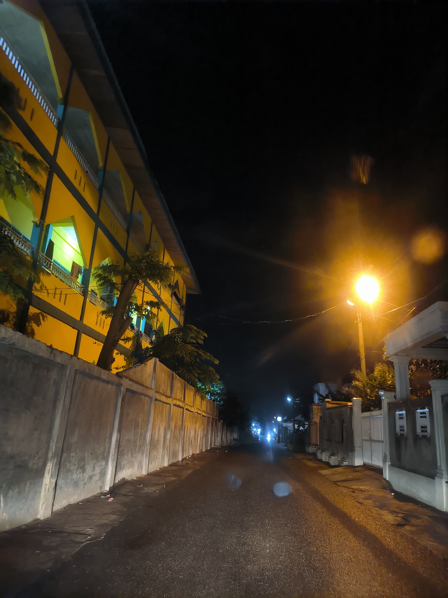 The image presents a nocturnal view of a quiet, narrow street in Lhokseumawe, Indonesia, after what appears to be a recent rainfall, as the asphalt road is visibly wet and highly reflective of light sources.
On the left, a prominent multi-story building with a bright yellow exterior and light blue accents runs along the street. This structure, possibly an institutional building or dormitory, features several levels with open corridors or balconies, some of which are illuminated by internal lights casting a greenish glow. A tall, textured concrete wall acts as a boundary between the building and the road. Trees with dark foliage are interspersed along this wall, with some branches extending upwards towards the building's facade.
On the right side of the street, a similar concrete wall defines the edge of what appears to be residential properties, leading to a gate structure with white pillars. A single, intensely bright streetlamp on a pole stands out on this side, casting a strong, warm orange light that dominates the right half of the scene and creates significant lens flare. Overhead wires are visible, crossing the street.
Further down the road, additional streetlights punctuate the darkness, and the faint headlights of a vehicle can be seen approaching, indicating minimal activity. The sky above is dark, contrasting sharply with the artificial lights that define the visible elements of the street. No people are discernible in the scene.