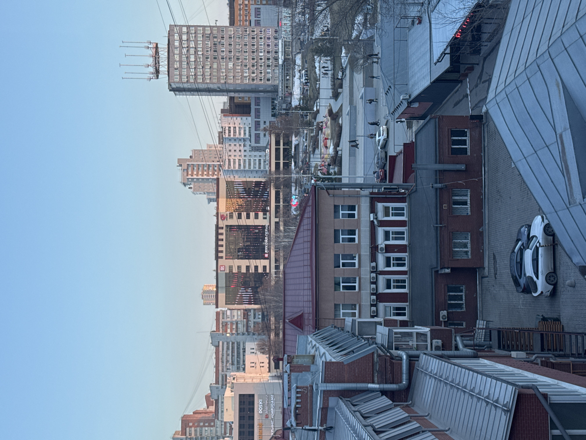 The image shows a view of Perm, Russia. It is daytime, with a clear sky. The scene includes several buildings, including high-rise apartments, office buildings, and older brick structures with metal roofing. Air conditioning units are visible on the sides of some buildings. In the background, there's a pedestrian area with people walking. A white car is parked on a brick-paved area. Power lines run across the view. The architecture and signage indicate the location is Perm, Russia.