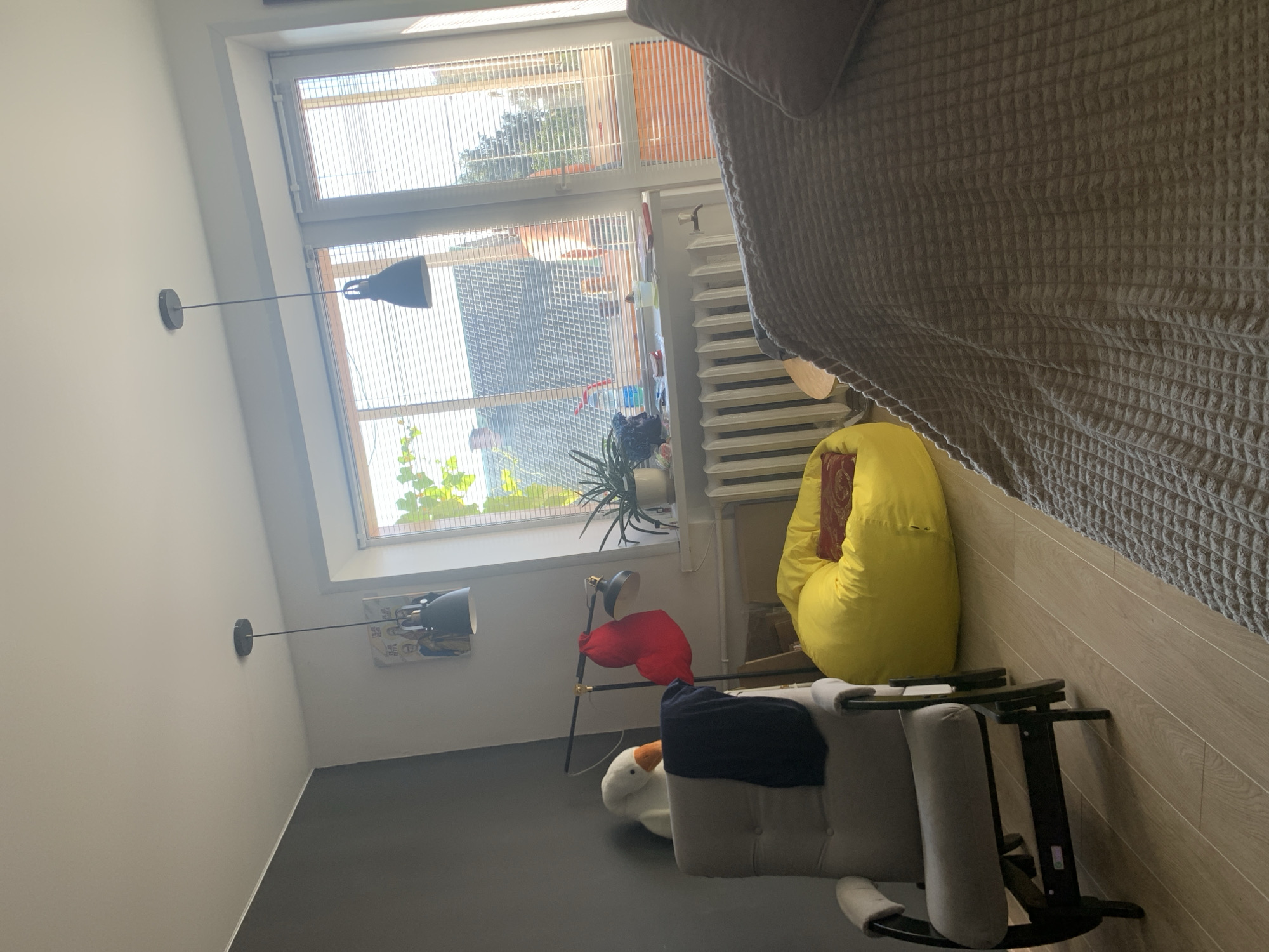 The image depicts an interior scene of a room, possibly a living area or bedroom, with natural light streaming through a large window. The window is covered by horizontal blinds and offers a view of residential rooftops and trees. Prominently featured are two hanging pendant lamps with dark conical shades. In the foreground, a section of a large, textured grey blanket or bedspread covers a surface, suggesting a bed or sofa. To the left, a light-colored upholstered glider chair with dark wooden accents is positioned. A dark navy fabric is draped over the back of the chair, and a white plush toy goose with an orange beak sits on the chair's armrest. Adjacent to the chair, a floor lamp with a dark shade is visible, with a bright red fabric item hanging from it. Further to the right, a large, vibrant yellow beanbag chair is placed on the wooden floor. A potted plant sits on the windowsill. The overall impression is one of a lived-in, cozy domestic space. The location is identified as the city of Kapyĺ, Belarus.