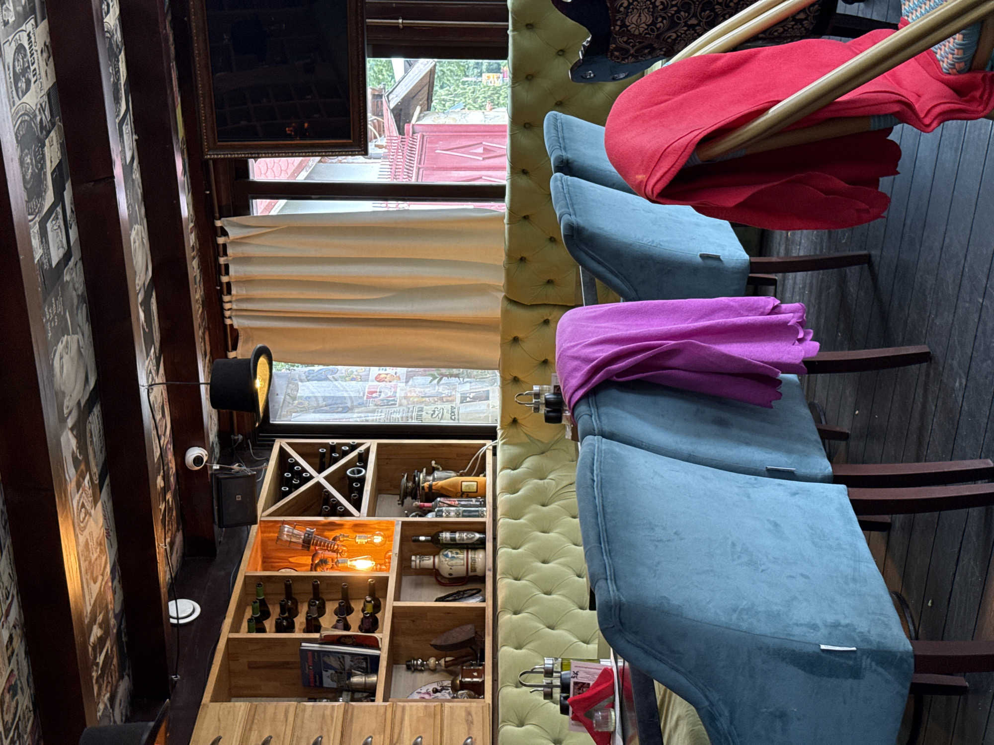 This media file depicts an indoor scene, likely a bar or restaurant, with a rustic and eclectic decor. The prominent subjects are several chairs upholstered in light green tufted fabric, some of which have a blue fabric draped over them. A red fabric is also draped over one of the chairs. To the left, a wooden shelf unit is filled with various objects. These include rows of wine bottles in the top section, decorative light bulbs and bottles in the middle sections, and what appear to be antique items like a sewing machine and an iron in the lower sections. A black top hat sits on a stand to the left of the shelves, near a security camera. The background reveals a window with a view of an outdoor area with red architecture and greenery. The overall ambiance suggests a cozy and possibly vintage setting, possibly in the evening or an interior space with artificial lighting. There is no explicit indication of an activity or event taking place, and no clear emotional cues are conveyed. The text visible on the wallpaper on the far left is fragmented and difficult to read, but appears to be in a newspaper-like style with images and headlines. The location context of Predeluț, Romania is provided.
