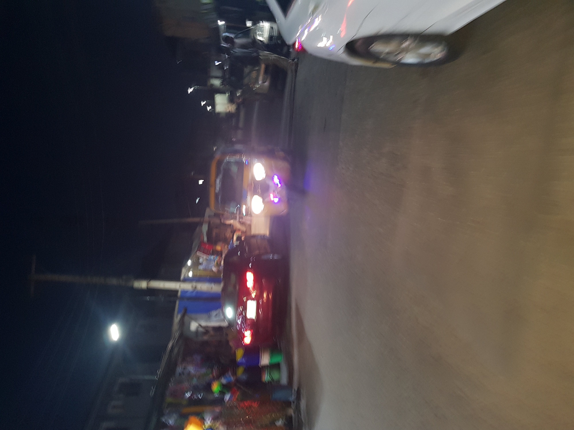 The scene shows a street in Shangisha, Nigeria, at night. The image is rotated to the left. A white car is partially visible at the top right. A yellow auto-rickshaw is further down the street, with its headlights on. Behind the auto-rickshaw, there is a dark car with illuminated taillights. A street vendor is positioned on the left side of the street, selling items. A street light is visible in the background. People are present on the side of the road.