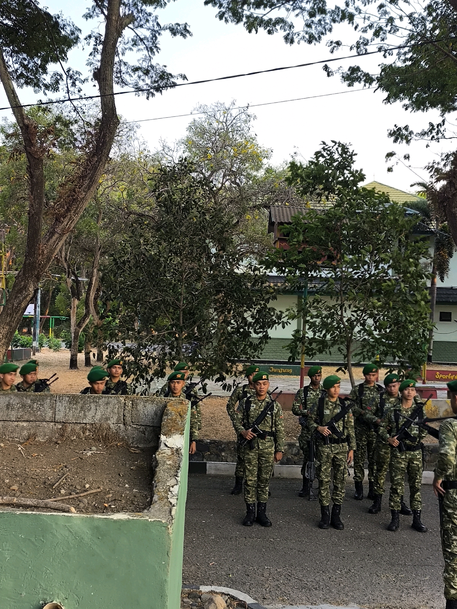 In this eye-level, medium shot, a group of approximately ten young Indonesian soldiers in camouflage uniforms and green berets stand at attention on a paved surface. They are holding rifles. Behind them, to the left, is a low, green-painted concrete wall filled with dirt and debris, with some branches and sticks visible. Beyond the wall, trees with dense foliage are present. In the background, there is a building with a yellow roof and visible windows. Signs with text such as "JUMJUNG" and "Sportif" are present on the building. The overall environment appears to be an outdoor training or parade ground in Indonesia.