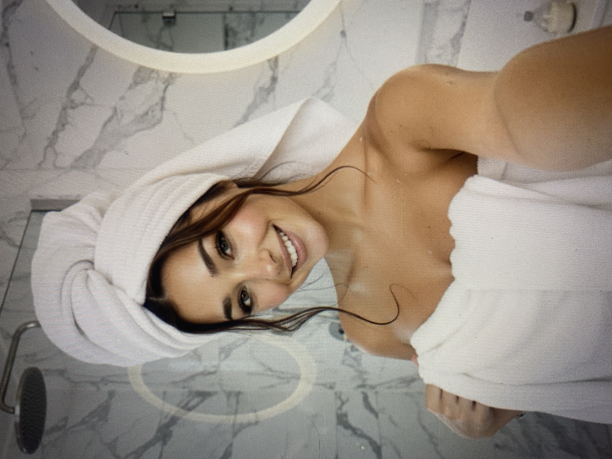 A young woman with a towel wrapped around her head and body is standing in a bathroom, holding another towel. She is smiling and looking at the camera. The bathroom has marble walls and a large, lit mirror.
