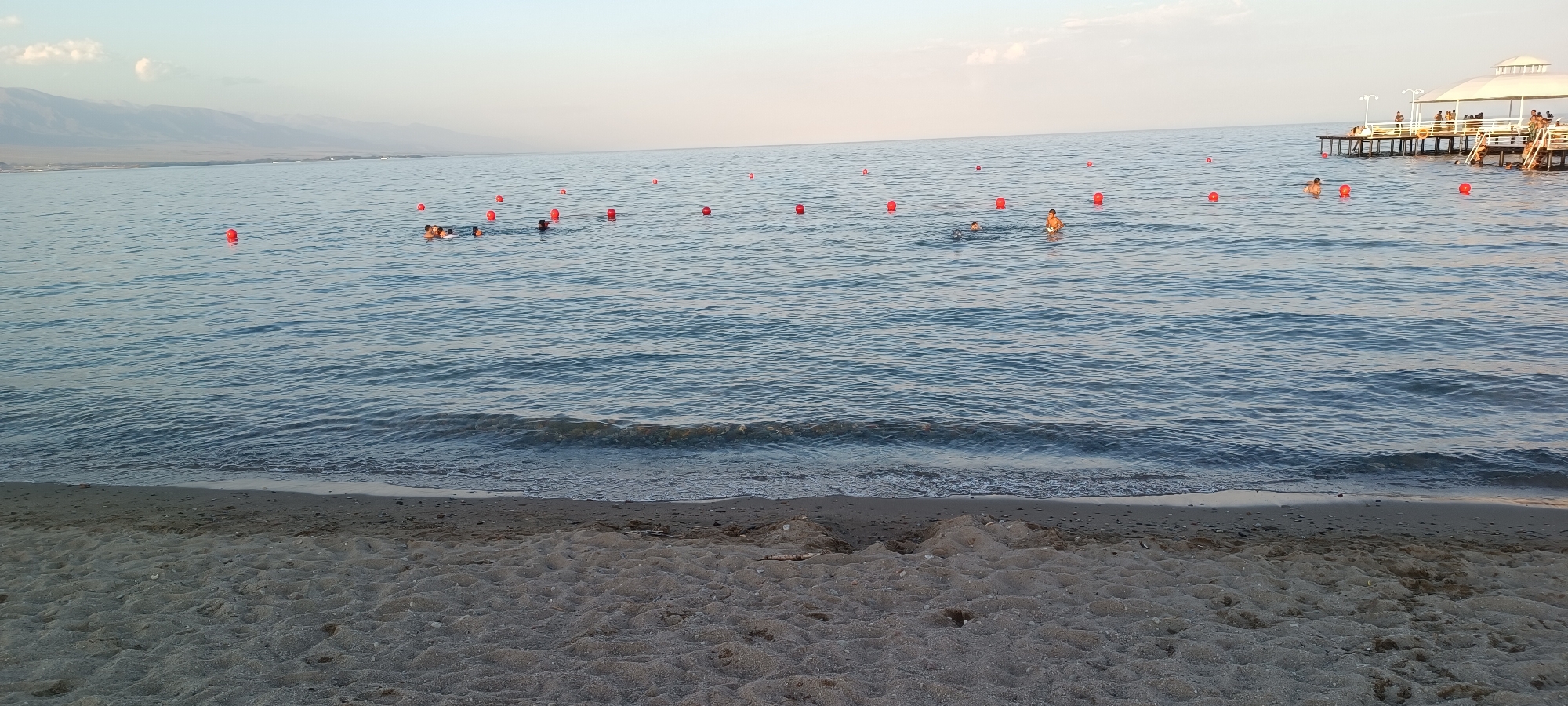 In Koshkol', Kyrgyzstan, this image captures a serene beach scene on a clear day. The foreground is dominated by a sandy shore with subtle footprints, leading to the gentle waves of a calm, blue body of water. In the water, several red buoys are scattered, marking a designated swimming area. A group of people are visible swimming and playing in the water, some near the buoys and others further out. In the distance, a wooden pier extends into the water, featuring a covered structure and several individuals relaxing on it. Beyond the water, a range of hazy, pale mountains forms the horizon, under a soft, pale blue sky with a few wispy clouds. The overall atmosphere is one of peaceful recreation.