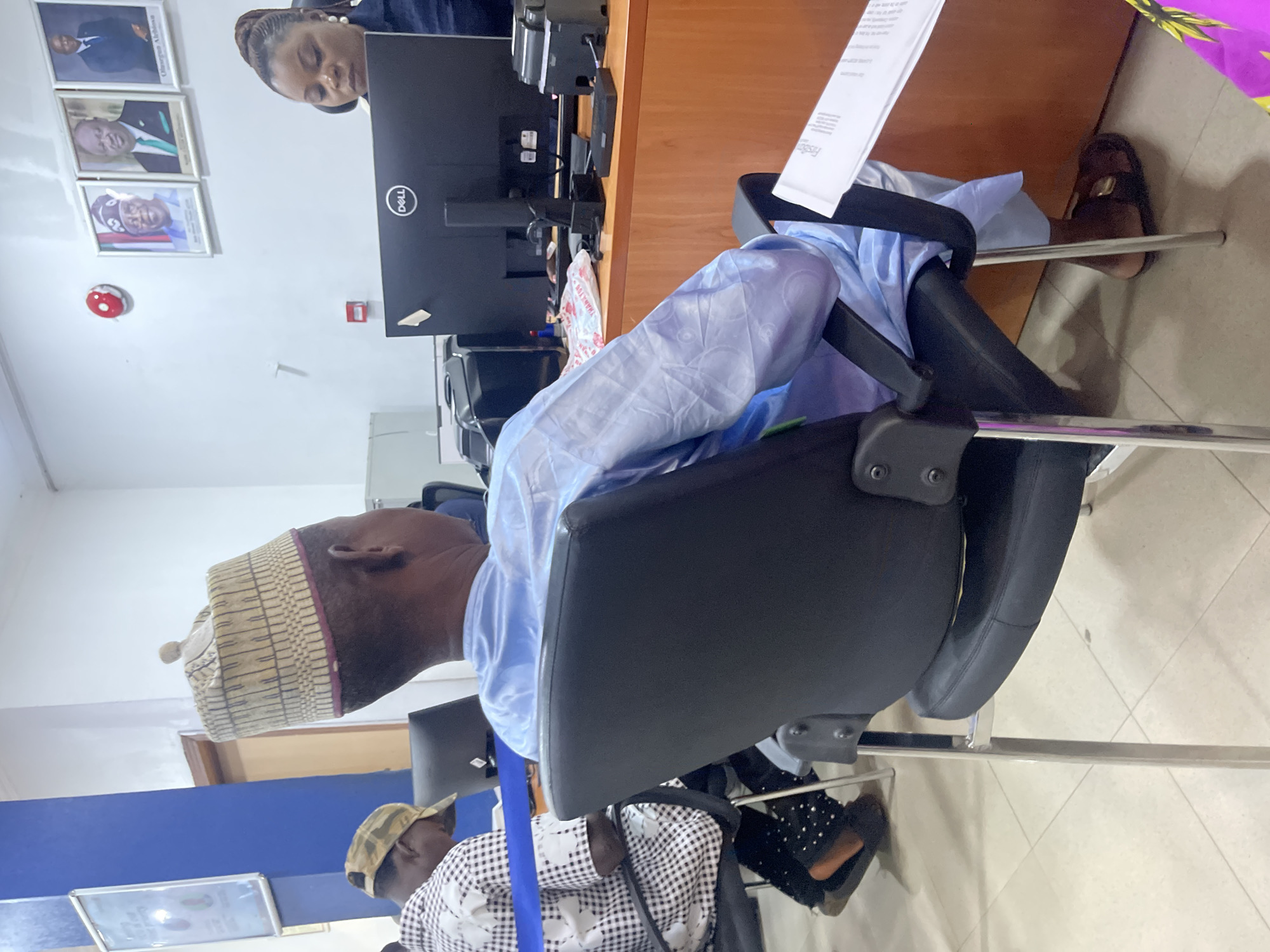 A man in a light blue traditional outfit and a patterned cap sits in a black office chair, facing away from the camera. He is positioned in the foreground to the right of the frame. To his left, another individual in a checkered shirt and a camouflage cap is also seated, facing away and slightly to the left. In the background, a woman in dark attire is visible behind a desk, looking at a computer monitor. The office environment includes a wooden desk with a Dell computer monitor, a printer, and various papers. Framed portraits of three men are mounted on the wall behind the desk. A blue stripe of tape is visible on the desk surface in front of the man in the checkered shirt. The floor is tiled with a beige and white checkered pattern. The overall scene appears to be a waiting area or service point within an office building. The presence of framed portraits of political figures and the Dell computer setup suggests an official or governmental setting, potentially within Jos, Nigeria.