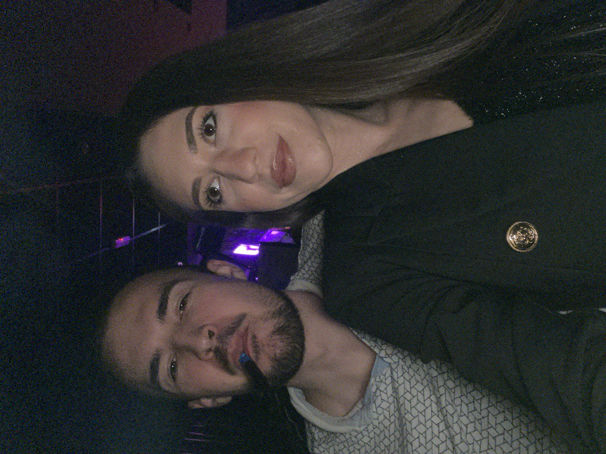 A photograph depicts a man and a woman in an indoor environment, possibly a nightclub or lounge, given the low light conditions. The woman has long brown hair and fair skin. She wears dark clothing with a visible gold button. The man, positioned lower in the frame, has short dark hair, a trimmed beard, and is wearing a patterned shirt with what appears to be a blue mouth guard or chewing gum. The background is dark with vague hints of interior structures. The location cannot be definitively identified as Sofia, Bulgaria based on visual information alone. Further information is needed to confirm the city location.