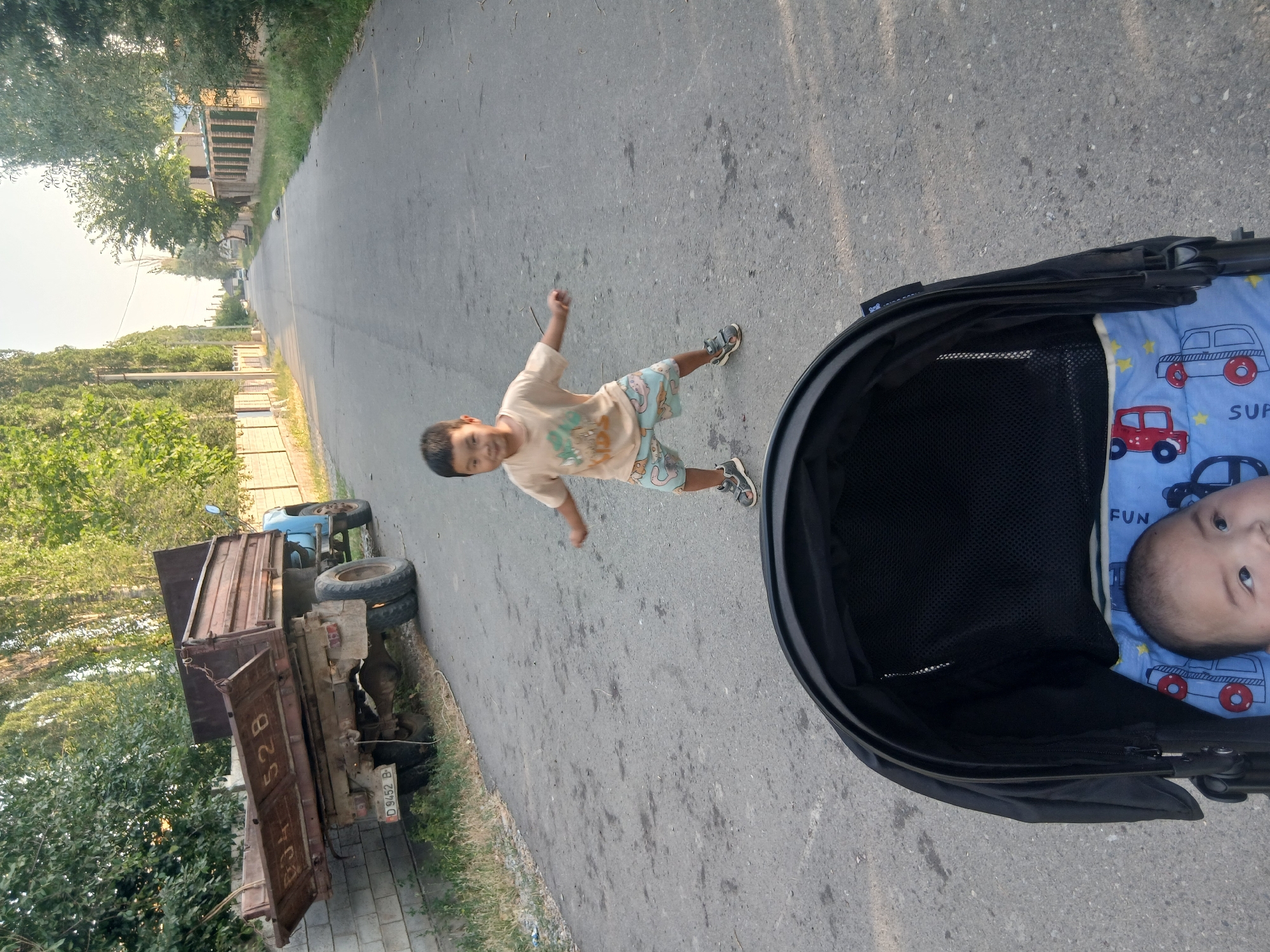 In the city of Taygarayev, Kyrgyzstan, a young boy is captured in motion on a paved road. He is wearing a light-colored t-shirt and patterned shorts, with sandals on his feet. The boy's stance suggests he is mid-action, possibly dancing or playing. In the foreground, a portion of a black stroller is visible, with a baby lying inside, adorned in a blue blanket with car and star patterns. In the background, a rusty, light blue truck is parked on the side of the road, leaning against a brick wall. The environment appears to be a residential street lined with trees and fences. The sky is clear and bright, indicating daytime.