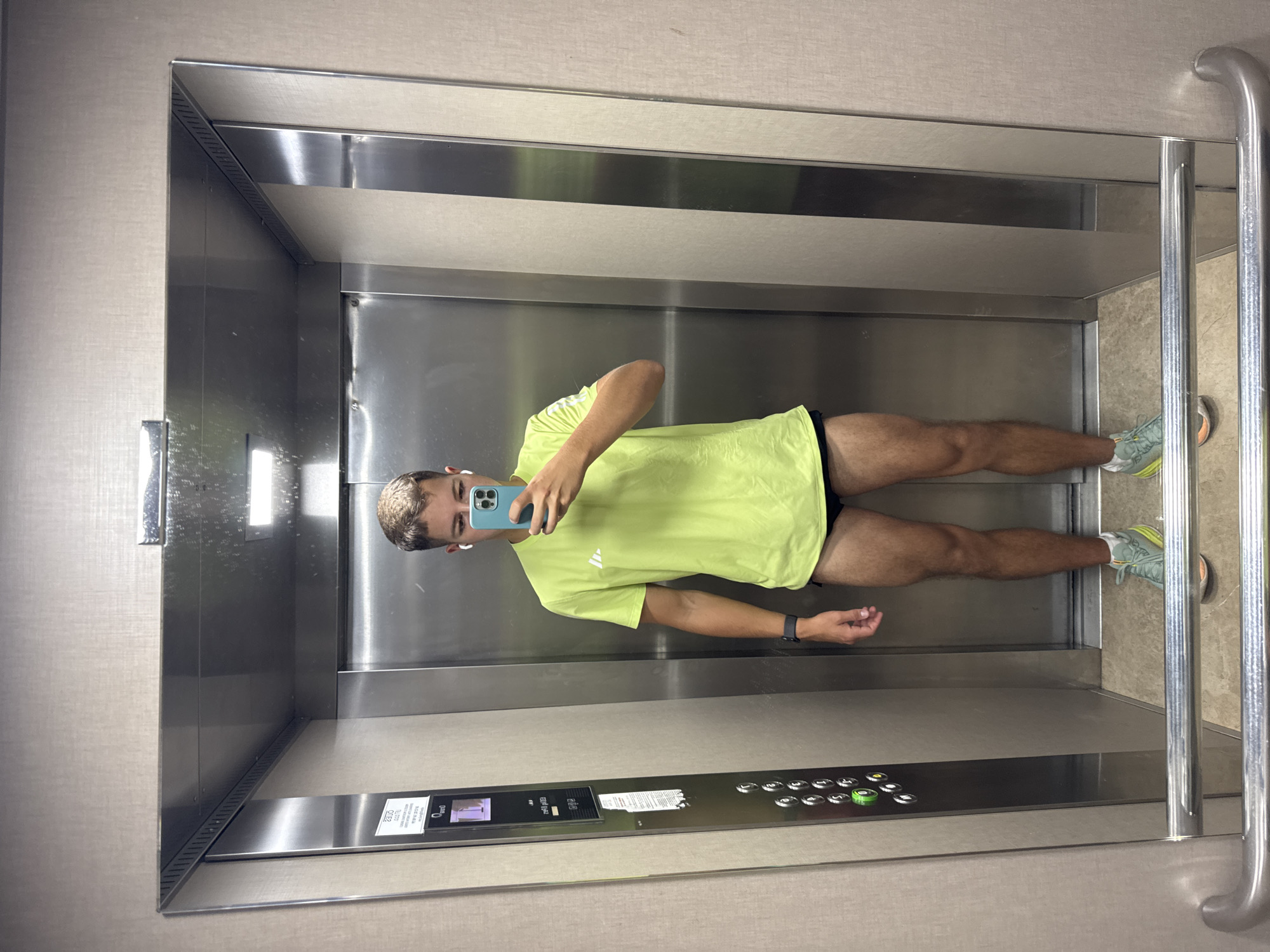 A young man with blonde hair stands in an elevator, taking a mirror selfie with his phone. He is wearing a bright green t-shirt with white stripes on the sleeves and black athletic shorts. He has on white earbuds and a dark smartwatch on his left wrist. His right hand holds a light blue phone. The elevator appears to be in a building with metallic walls and a patterned beige wall. There are elevator buttons visible on the left side of the elevator. The floor of the elevator is tiled and there is a metal handrail at the bottom.