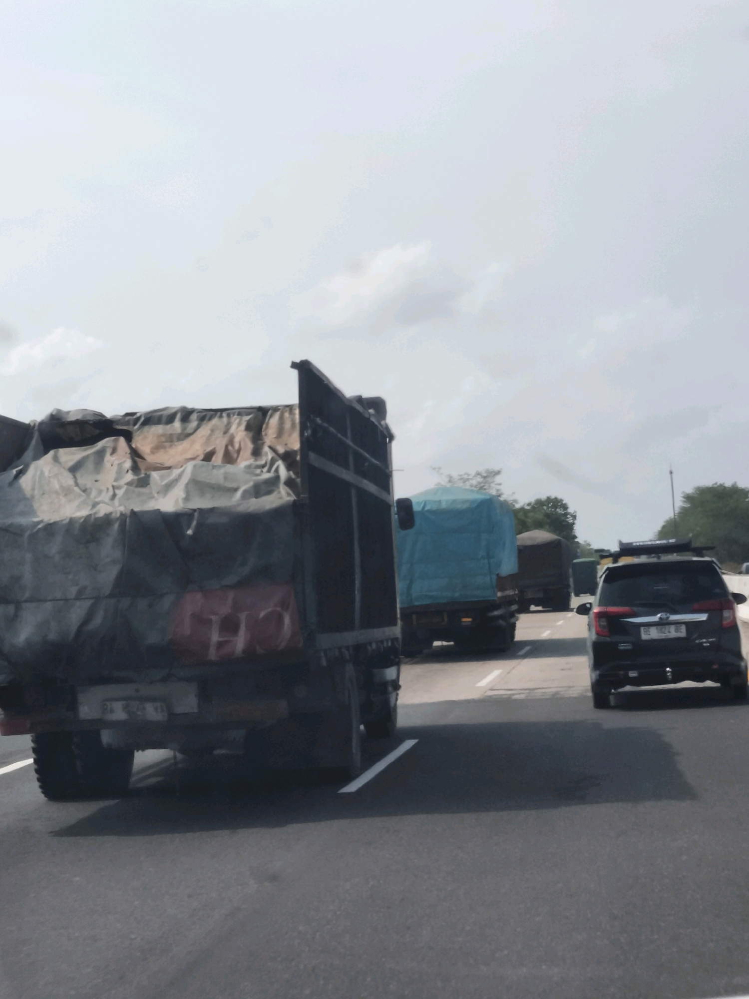 This image captures a view of a highway with several vehicles on a cloudy day, likely in Lubuk Dalam, Indonesia. The foreground is dominated by the rear of a large truck covered with a tarp that appears to be a patchwork of different colored materials, some with red markings. The truck has large rear tires and a visible license plate. To the right of this truck, a black SUV is visible in the next lane, also with its rear facing the camera, displaying a license plate. Further ahead on the highway, another truck with a bright blue tarp is visible, and beyond that, other vehicles and trees line the side of the road. The road surface is asphalt with white lane markings. The sky is overcast with diffuse clouds.