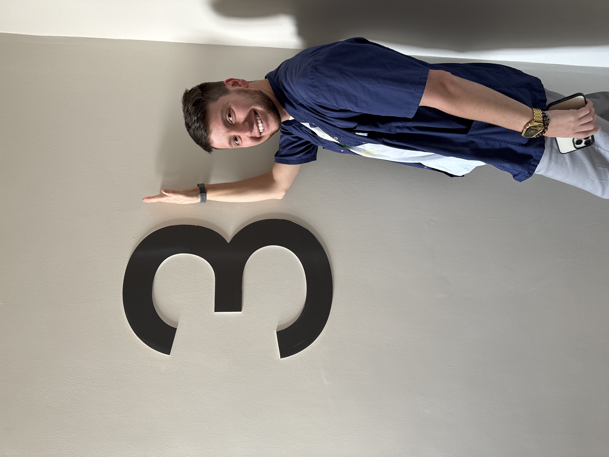 A young man with dark hair and a beard smiles at the camera while leaning against a light-colored wall. He is wearing a navy blue scrub top over a white shirt and light grey pants. On his left wrist, he has a dark fitness tracker, and on his right wrist, he wears a gold watch and beaded bracelets. He is holding a white smartphone in his right hand. To his left, a large, black, three-dimensional numeral "3" is mounted on the wall. The scene appears to be indoors, likely in a hallway or examination room. There are no discernible features that would indicate a specific location within Moscow, Russia.
