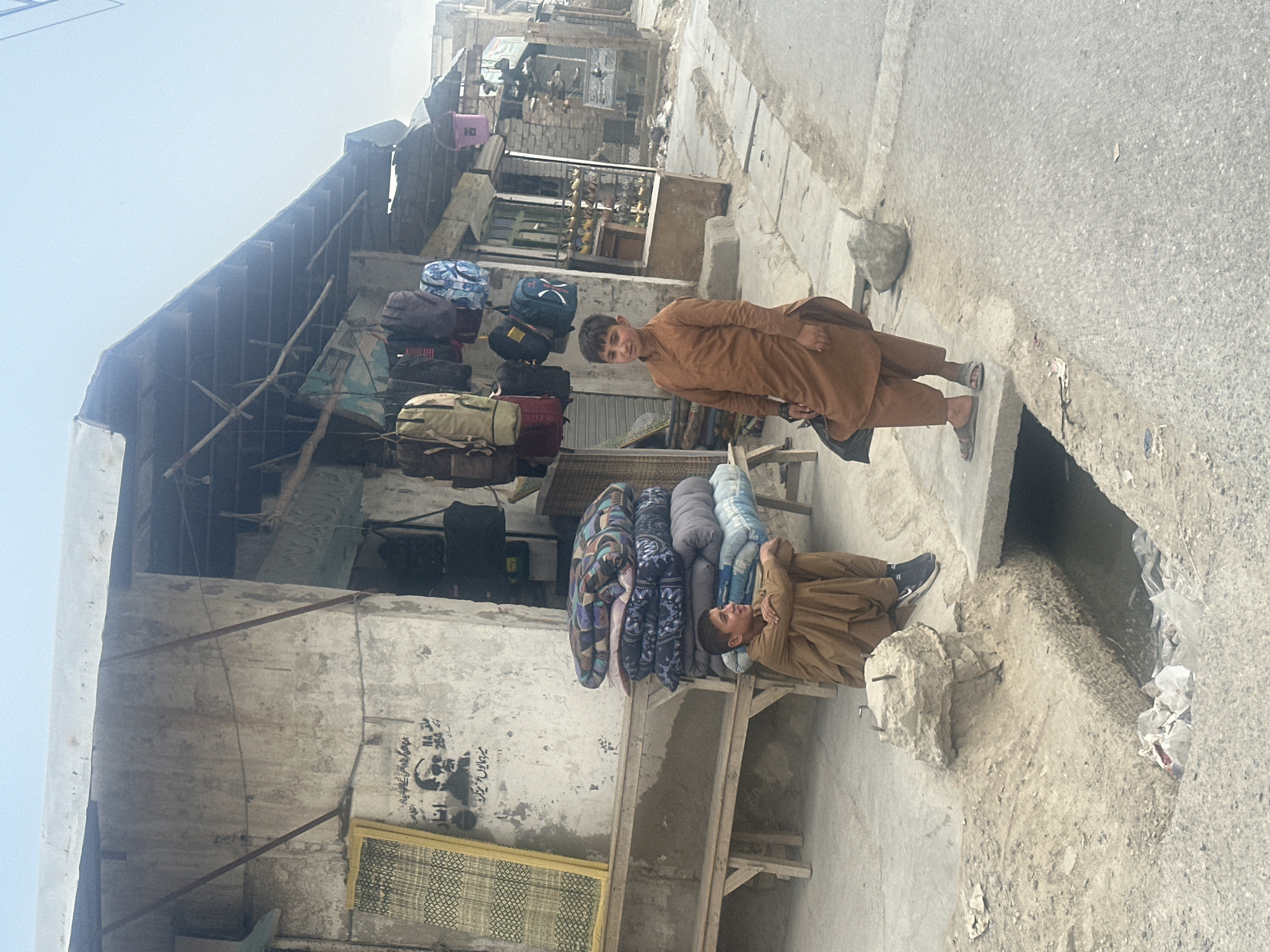 The image depicts a street scene in Quetta City Tehsil, Pakistan. Two boys are present. One is standing near a street, wearing a brown outfit. The other boy is sitting on a wooden platform, leaning against a stack of quilted blankets. Backpacks are displayed above the wooden platform. The surrounding buildings appear weathered, with one showing graffiti. The road is paved, with litter visible in the gutter.