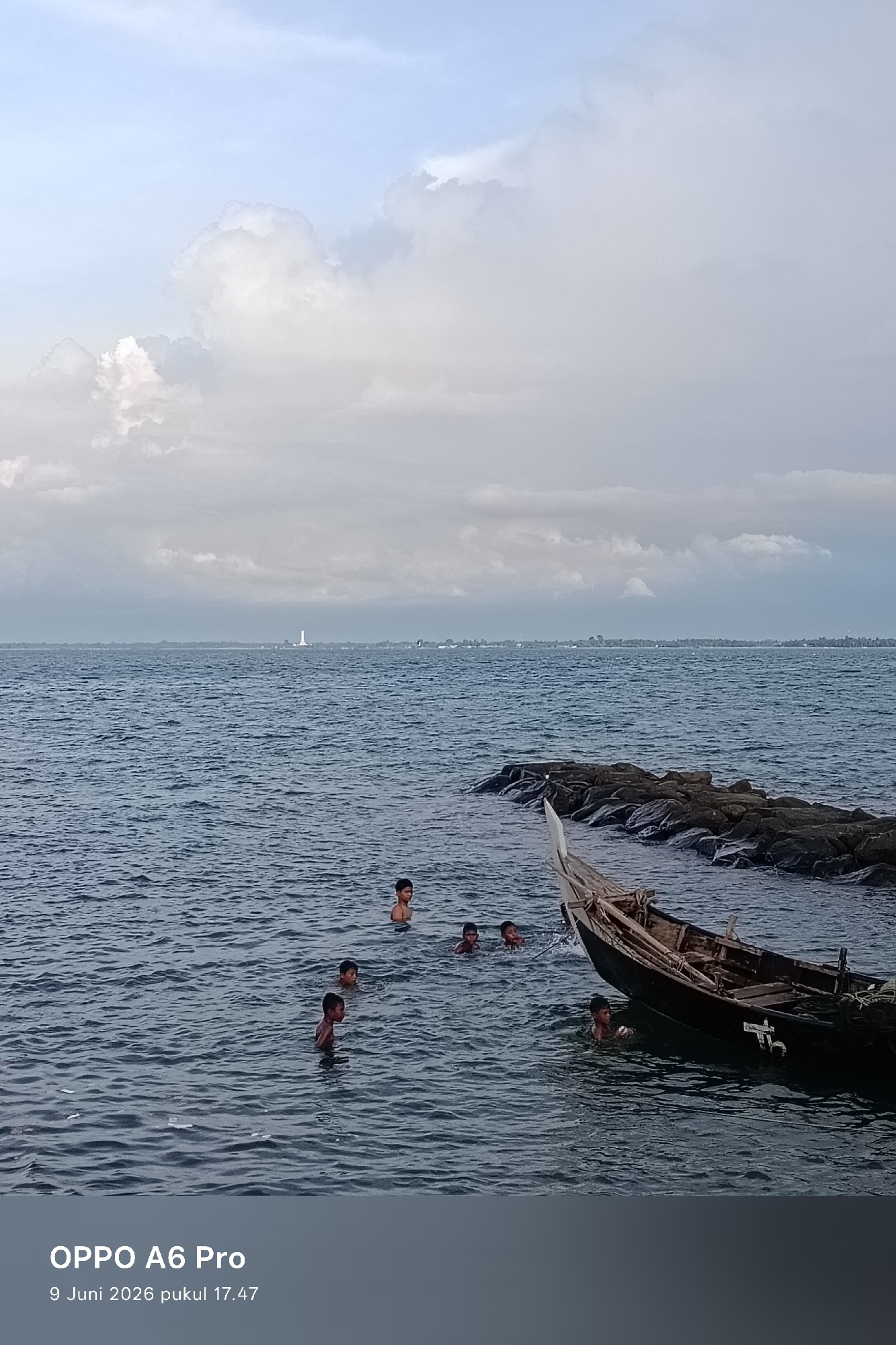 In Lhokseumawe, Indonesia, children swim in the dark blue ocean. A traditional wooden boat is moored near a breakwater made of rocks. The sky is filled with a mix of white and grey clouds, with a hint of blue peeking through. In the distance, a lighthouse and a distant shoreline are visible. The timestamp "OPPO A6 Pro 9 Juni 2026 pukul 17.47" is overlaid on the image.
