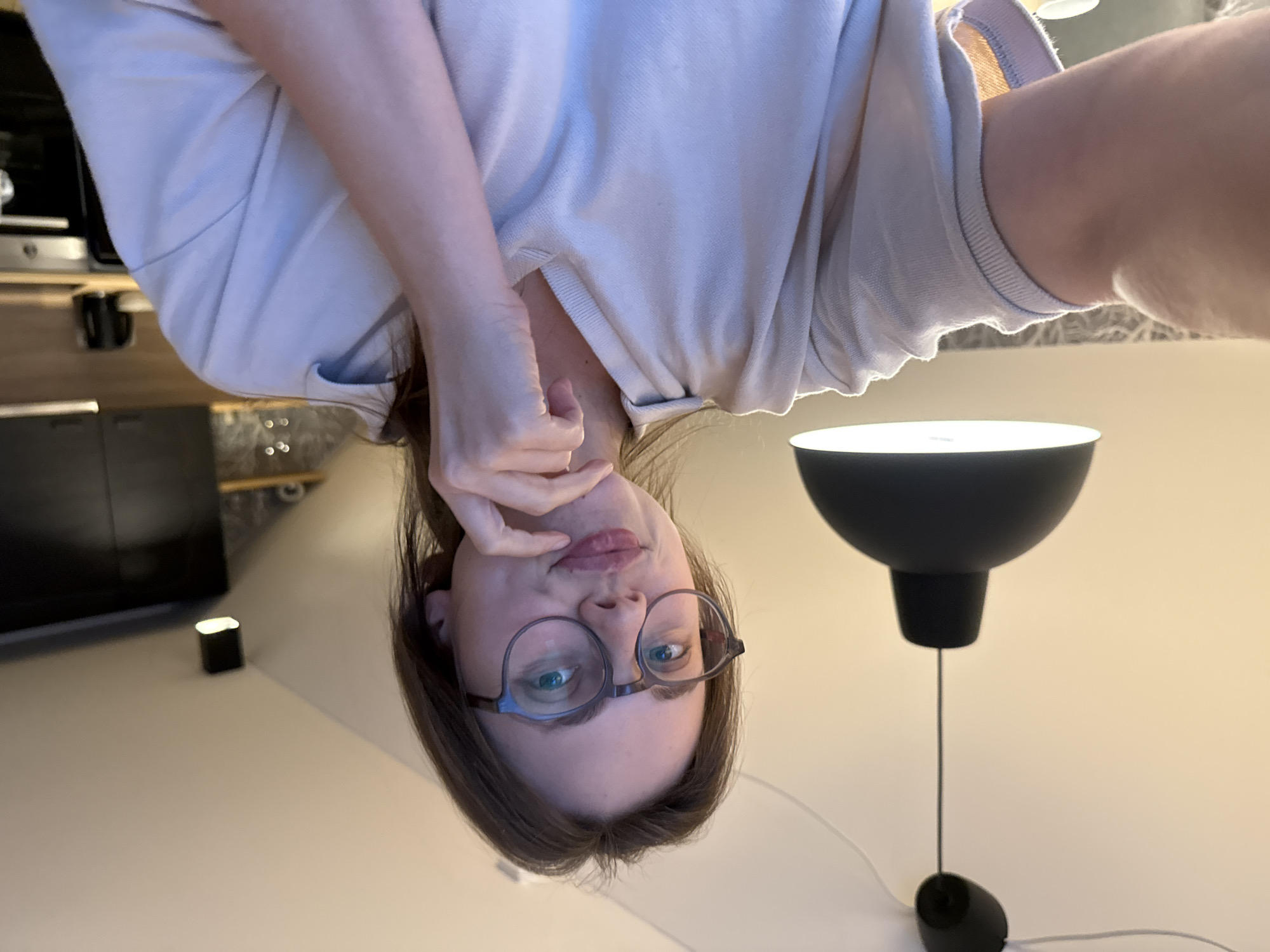 A young woman with brown hair and glasses is looking at the camera, touching her chin with her hand. She is wearing a light-colored polo shirt. The room appears to be a kitchen with dark cabinets and a black kettle on the counter. A black pendant light hangs from the ceiling. The scene is indoors, and the location in Vrchlabí, Czechia, cannot be determined from the visual information.