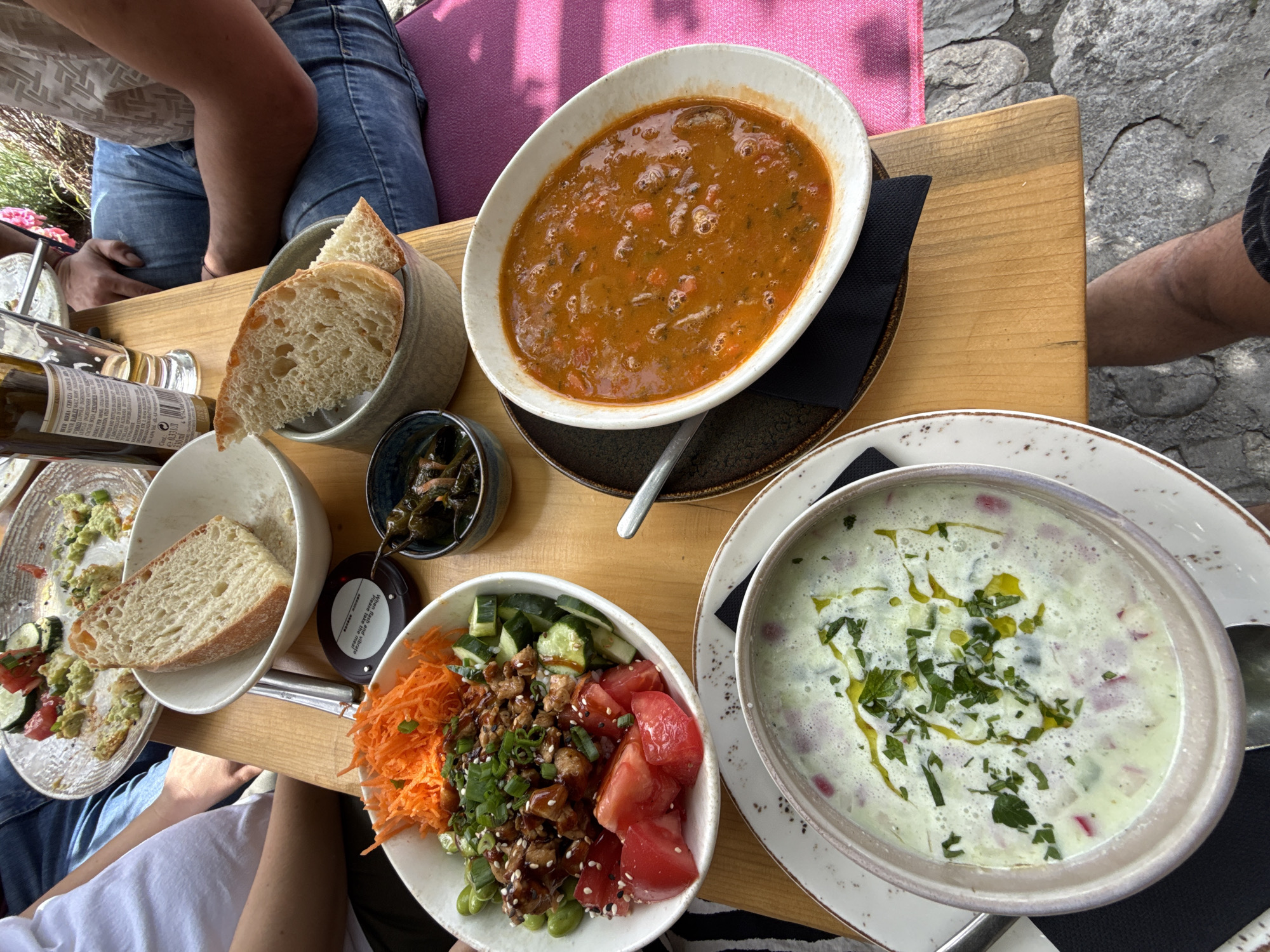 In Bansko, Bulgaria, a wooden table holds an assortment of food items, suggesting a meal shared outdoors. At the center, a white bowl contains a vibrant salad with diced tomatoes, cucumber, shredded carrots, and edamame, topped with chopped green onions and sesame seeds, drizzled with a dark sauce. To its left, a slice of crusty bread rests in a light gray bowl. Adjacent to the salad, a dark bowl holds what appear to be pickled jalapeños. Further to the right, a white, oval-shaped bowl is filled with a hearty, reddish-orange stew, likely a lentil or vegetable soup with chunks of carrots and other ingredients. Below this, a portion of a pink cushion is visible, indicating outdoor seating. In the foreground, another white bowl, this one with a speckled rim, contains a creamy, pale green soup, garnished with fresh herbs and olive oil, and studded with small red pieces, possibly radishes or beets. A spoon rests on the rim of this bowl. In the background, parts of two individuals are visible: one in a white t-shirt on the left, and another in blue jeans on the right, with an arm resting on the table. A beer bottle with a label is also present, along with part of a beer glass. The table rests on a stone-covered surface, suggesting an outdoor restaurant setting in Bansko.