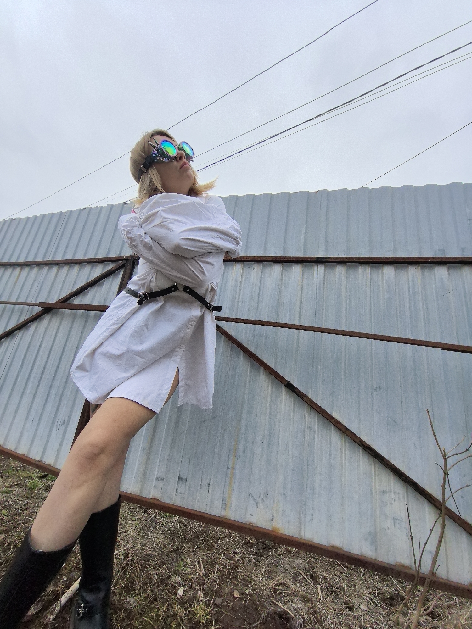 An outdoor, low-angle shot captures an adult female standing next to a tall, corrugated metal fence, potentially in Noviy, Russia. The woman, with blonde hair, is looking upwards towards an overcast, light grey sky, which features several dark power lines stretching across it. She is wearing striking iridescent or holographic goggles pushed onto her forehead, reflecting various colors. Her attire consists of a white, long-sleeved garment that resembles an oversized shirt or a stylized straight jacket, cinched at the waist by a black belt with buckles. Her arms appear to be crossed or held within the sleeves of the garment. She is also wearing black boots.
The background is dominated by the grey corrugated metal fence, which has rusty brown metal bars running horizontally and diagonally across its surface, providing structural support. The ground beneath the fence is covered with dry, sparse grass and a few bare tree branches. The scene conveys a somewhat gritty or industrial-edge setting under a cloudy daytime sky. No text is visible in the image.