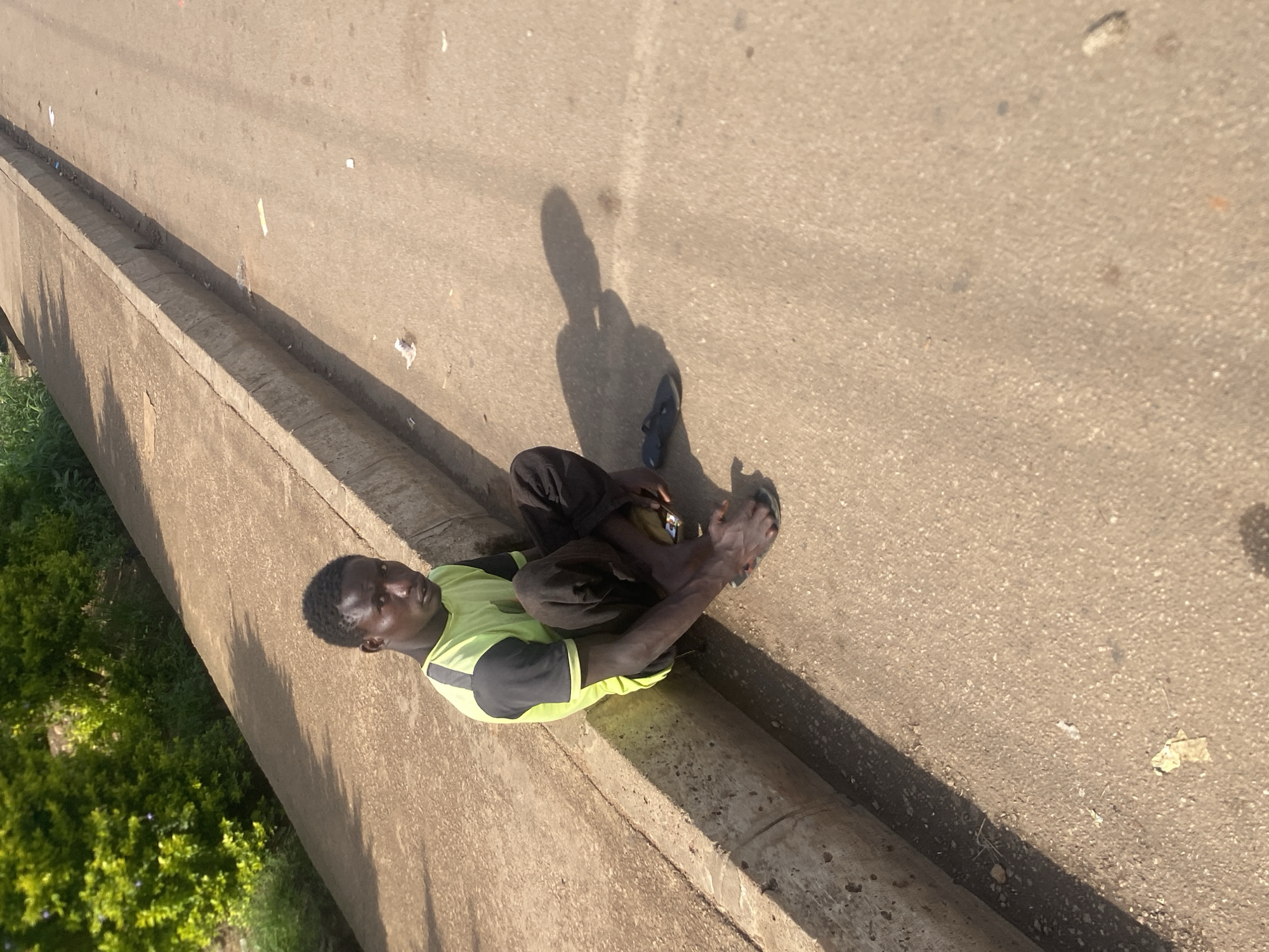 In Bukuru, Nigeria, a young Black male with dark skin and short, dark hair is seated on the ground next to a concrete curb. He is wearing a lime green t-shirt with black sleeves and black pants. His legs are pulled up, and he is looking towards the camera. A pair of dark-colored flip-flops are on the ground near his feet, and a shadow of his head is cast on the asphalt behind him. The ground is paved with asphalt and there is a concrete sidewalk or walkway to his left, bordered by a dense green bush. Scattered pieces of litter are visible on the ground.