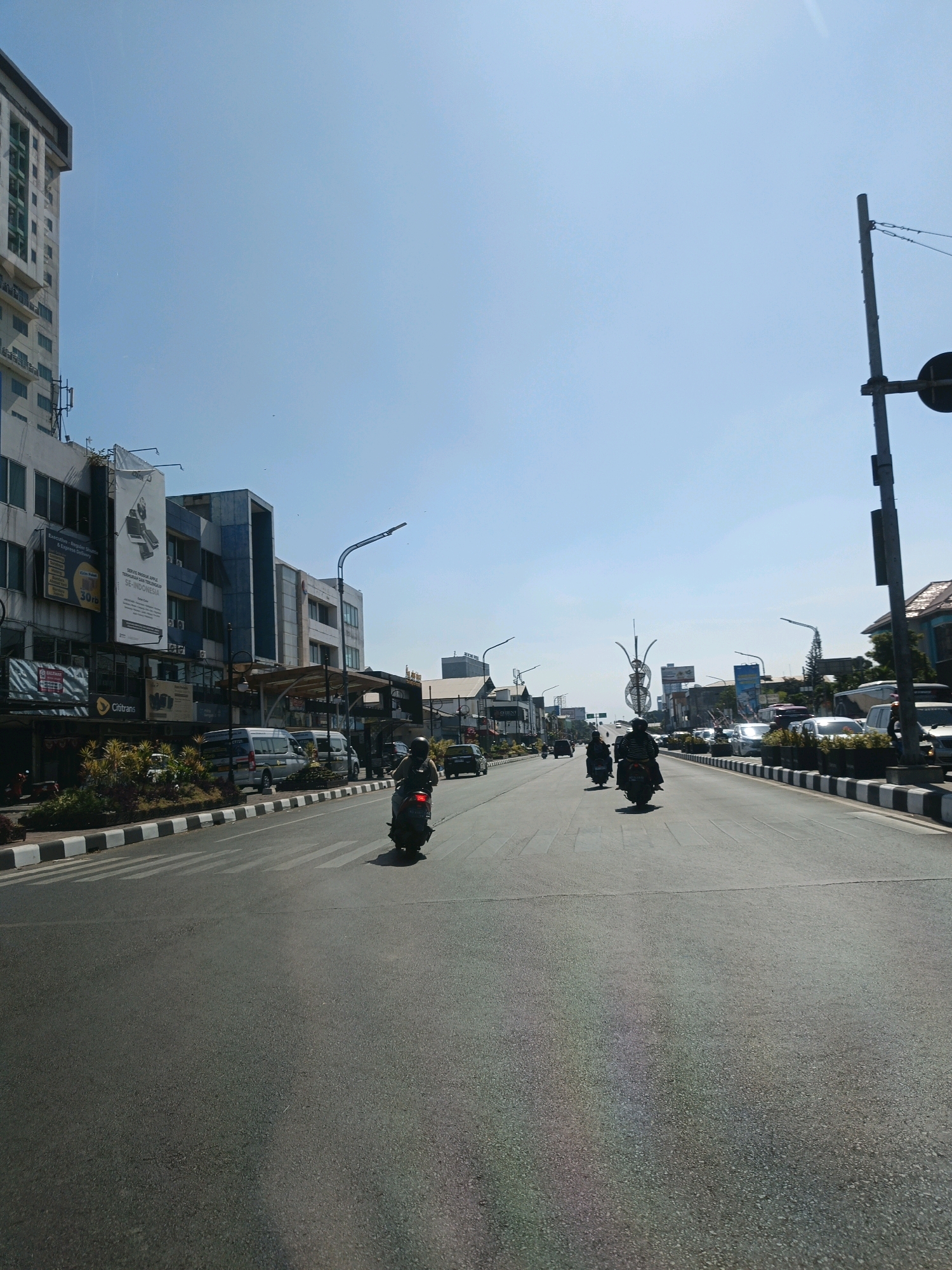 The image depicts a street scene in Surakarta, Indonesia, under a clear blue sky. The perspective is from a moving vehicle, looking forward down a multi-lane road.
On the left side of the road, there is a tall, modern building with multiple stories and glass windows. Adjacent to it are lower buildings with commercial signage, including a large banner for "Cititrans." Several parked vehicles, including silver vans, are visible along the roadside. There is a well-maintained green space with plants bordering the sidewalk, separated from the road by a striped curb.
The road itself is paved and appears to be a main thoroughfare. Several motorcycles are in motion, with one in the foreground to the left and two further down the road. Cars and other vehicles are also present, forming a line of traffic. Streetlights and traffic signals are visible.
On the right side of the road, there are more buildings, some with traditional-looking roofs. A tall, decorative metal structure stands on the right side of the road in the distance. The traffic appears to be moving, and the overall atmosphere suggests a daytime urban environment. The presence of multiple lanes of traffic and commercial buildings indicates a busy urban area.