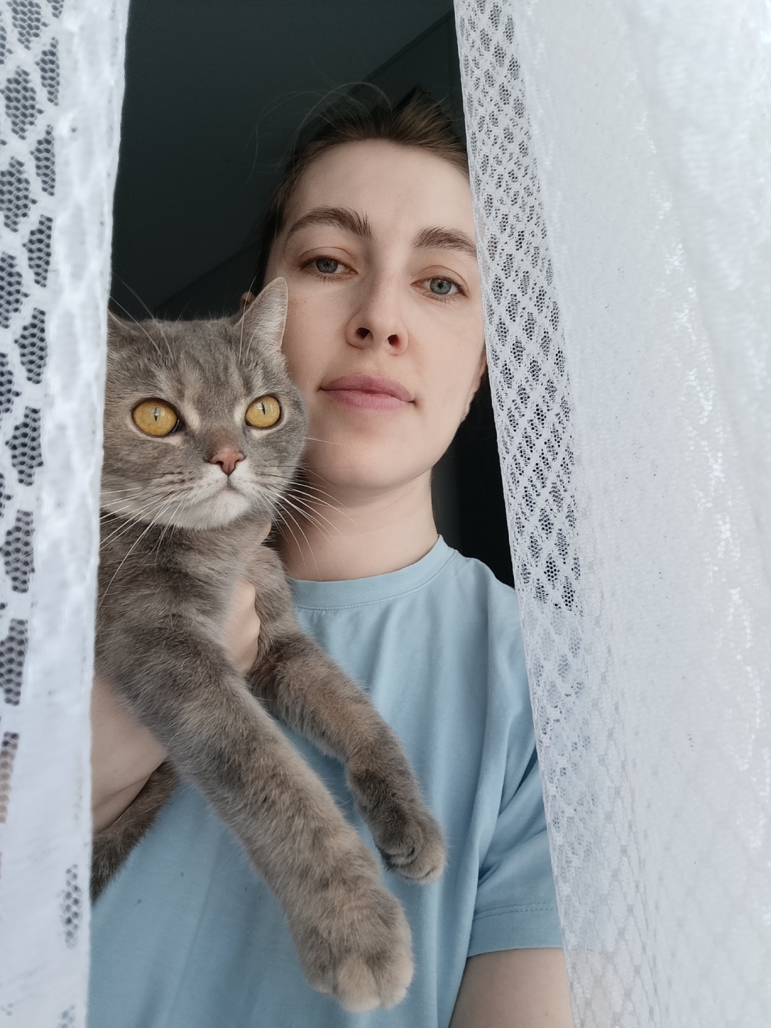 A woman is holding a gray tabby cat. They are near a window with white, lace curtains. The background is a dark wall. The woman has light skin and is wearing a light blue t-shirt. The cat has yellow eyes. No specific location or city is discernible from this image.