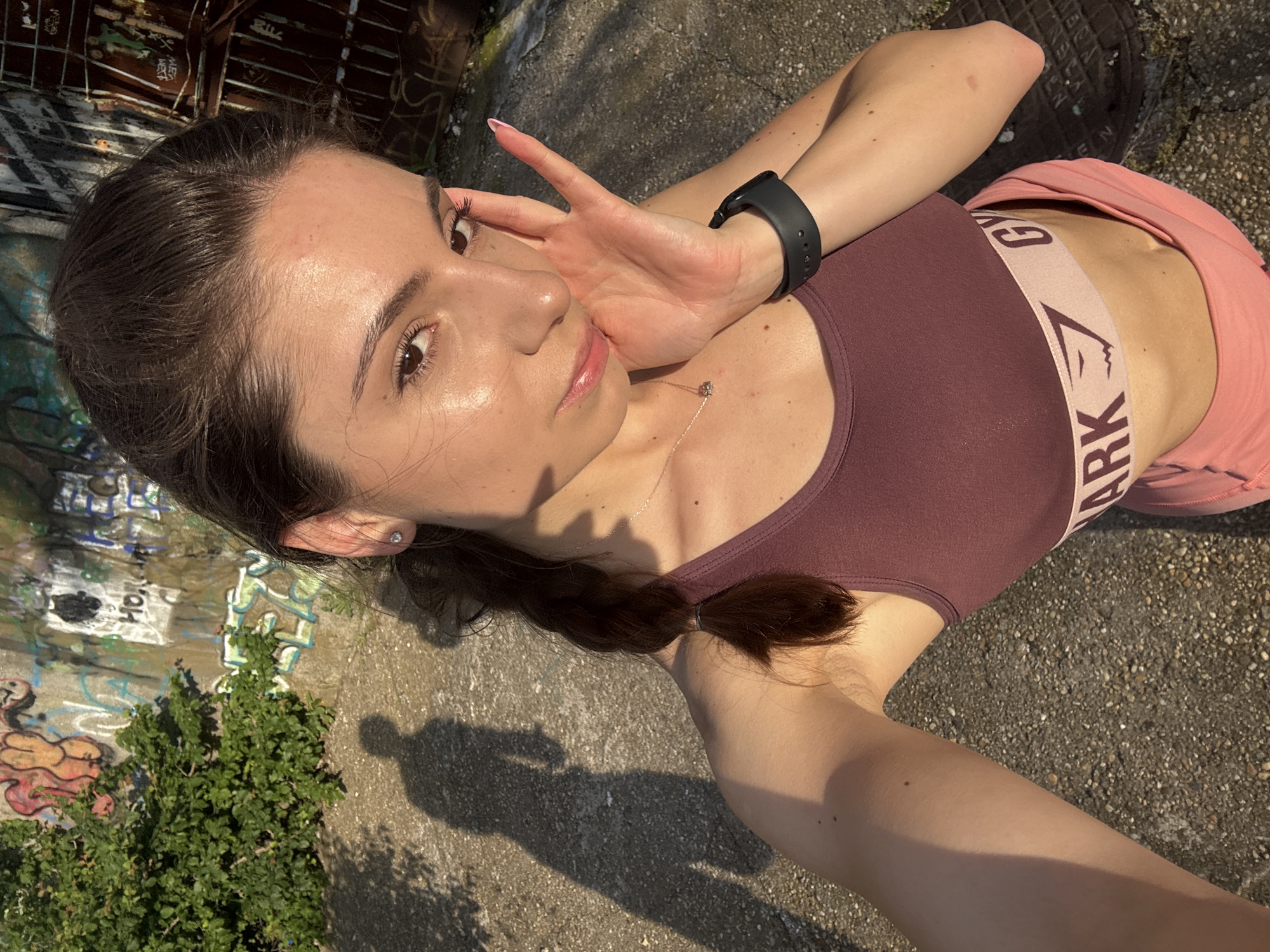 A young woman with dark hair in a braid, wearing a maroon sports bra and pink shorts, is taking a selfie outdoors. She is touching her face with her right hand, which has a black smartwatch on her wrist. Her shadow is visible on the ground to her left. The background shows a concrete wall covered in graffiti, some green foliage, and a rusty metal structure. The image appears to be taken in an urban environment, possibly an industrial or disused area. The lighting suggests it is either early morning or late afternoon, with the sun casting long shadows. The specific location in Varna, Bulgaria, cannot be determined from the visual information provided.