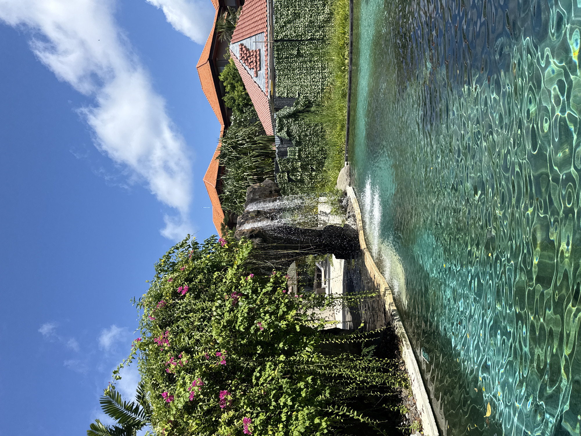 The photograph depicts an outdoor scene with a body of water, lush vegetation, and a building with orange tiled roofs, possibly in Unknown, Indonesia. The water has a turquoise hue and a rippled surface with reflections visible. A waterfall feature cascades into the water. A building with orange tiled roofs is situated amidst the greenery. The sky is visible in the background with blue sky and some white clouds. A bush with pink flowers is present in the foreground.