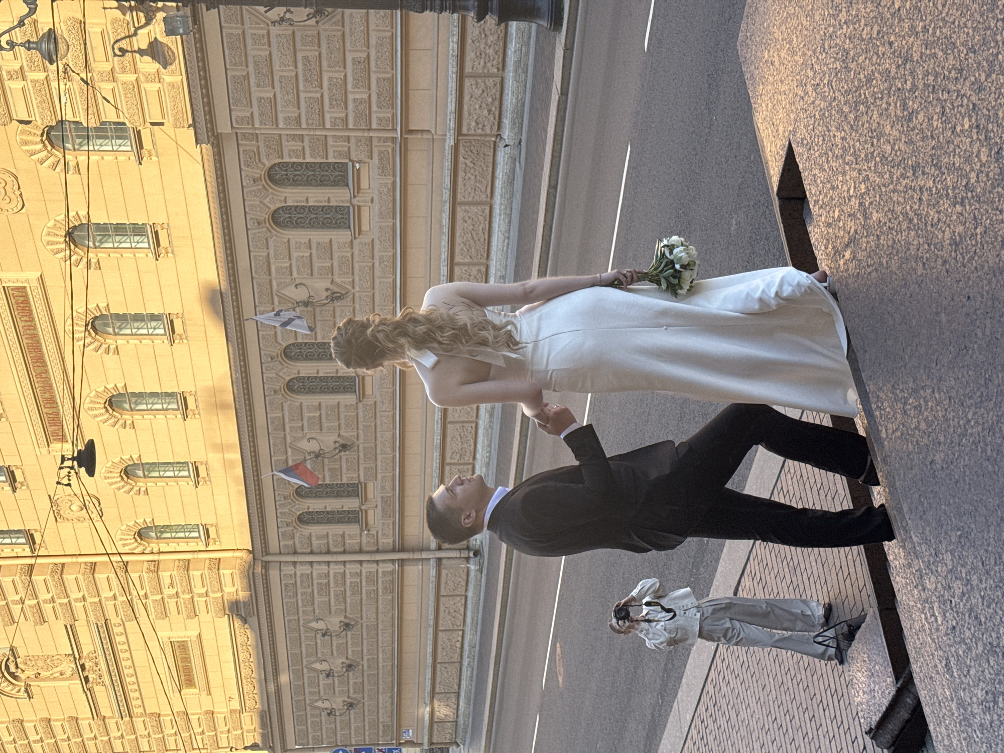 In Saint Petersburg, Russia, a couple, appearing to be newlyweds, is photographed on a sunny day with a backdrop of an ornate building. The man, dressed in a black suit and white shirt, holds the hand of the woman, who is wearing a white, strapless wedding gown and carrying a bouquet of white flowers. Their pose suggests they are engaged in a wedding photoshoot. In the foreground, a third individual, dressed in a white top and light-colored pants, is captured taking a photograph of the couple with a DSLR camera. The scene unfolds on a paved walkway next to a street, with a grand, light-colored building featuring arched windows and decorative stonework dominating the background. Russian flags are visible on the building's facade. The lighting indicates it is late afternoon or early evening, with long shadows cast by the setting sun.