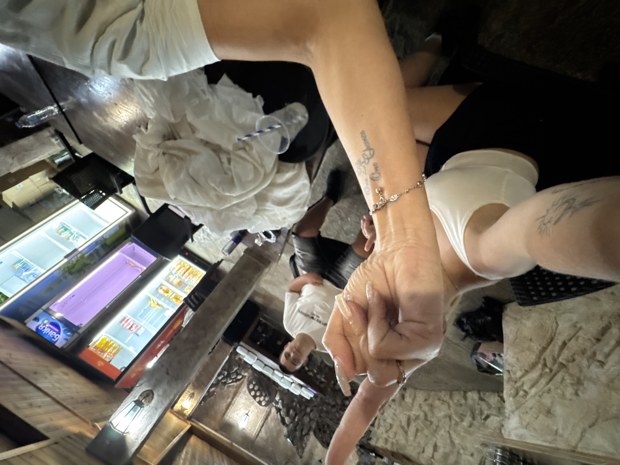 The image captures an indoor scene, possibly a bar or lounge, with a rustic ambiance. The flooring appears to be dark wood. In the background, a refrigerated display case is visible, stocked with beverages. A young man wearing a white t-shirt and dark shorts is seated, looking towards the camera. In the foreground, a person's arm and hand are prominent, with long, manicured fingernails adorned with glitter. A tattoo is visible on the arm, and a bracelet is worn on the wrist. A white piece of fabric, possibly clothing, is draped over a table. The lighting in the scene is dim, with artificial light sources contributing to the illumination. There is no explicit indication of the location being in Sredets, Bulgaria, or any other specific city.