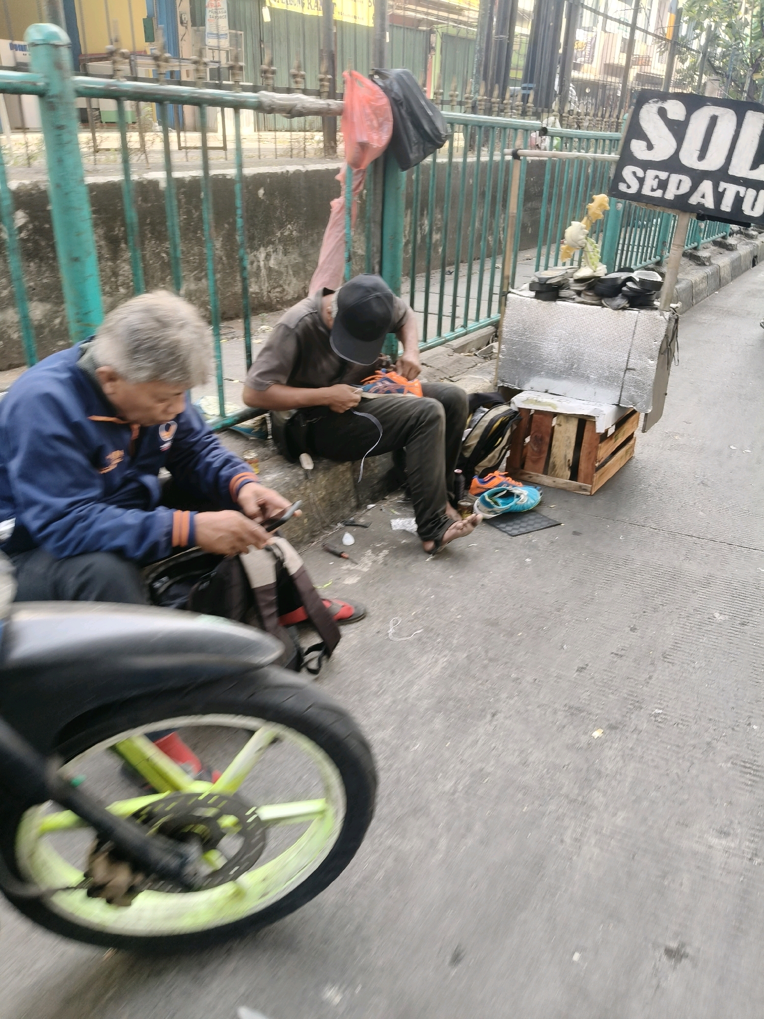 Two shoe repairmen are working on a sidewalk in front of a green metal fence. The shoe repairman on the left is wearing a blue jacket and black pants. He is using a smartphone and appears to be working on a shoe or sandal. The shoe repairman on the right is wearing a dark shirt and black pants. He is also working on a shoe and appears to be holding a tool. Next to him is a wooden cart with various shoe parts and tools on it. A sign that says "SOL SEPATU" (shoe soles in Indonesian) is also visible. The scene suggests a typical street-side business in South Tangerang, Indonesia. The sidewalk is paved, and there are some plants and debris on the ground. A motorcycle wheel is visible in the foreground.