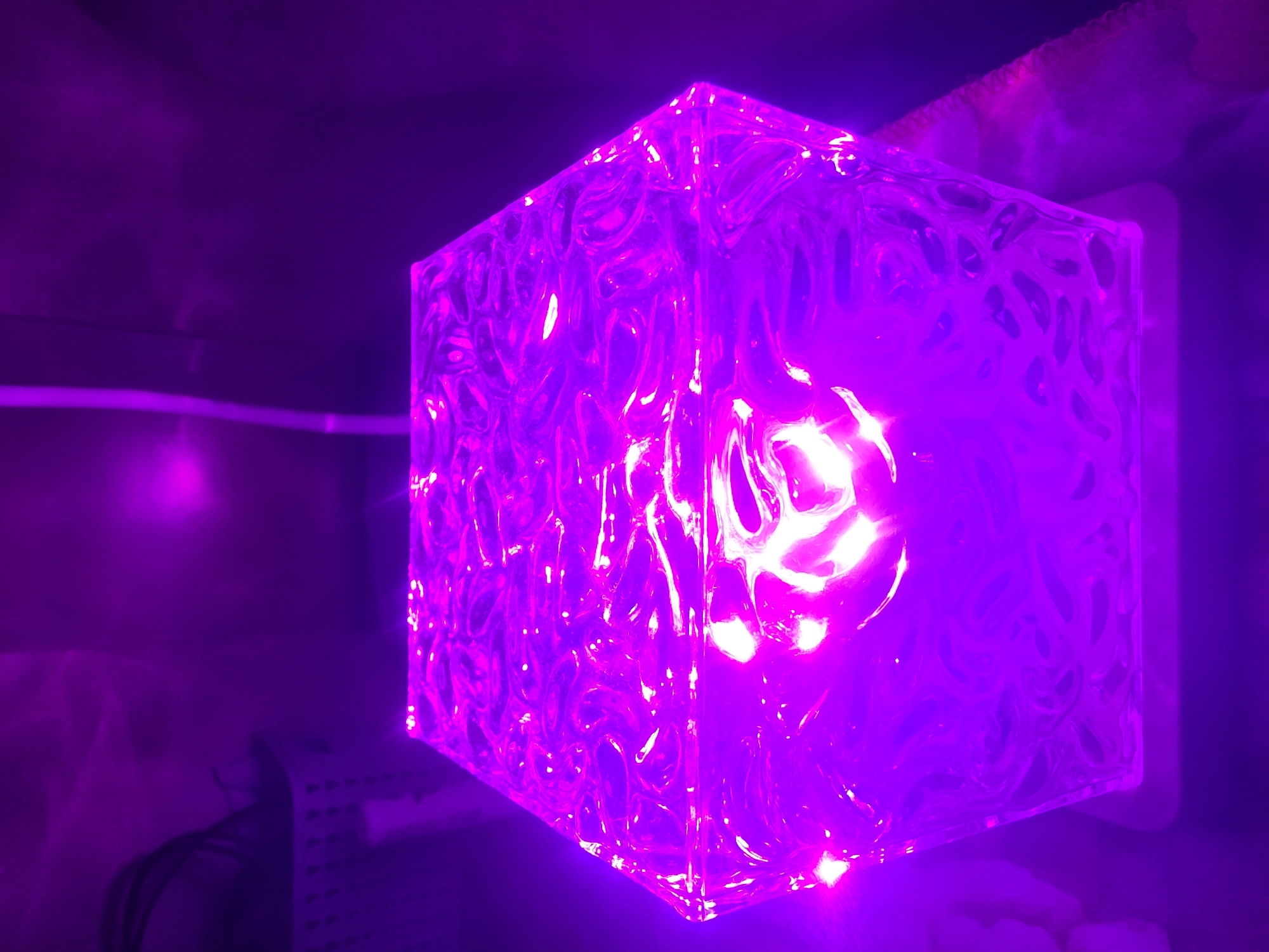 A hexagonal prism shaped object with a textured surface is illuminated from within by a bright pink light, casting a purple glow on its surroundings. The object rests on a white base, which is slightly visible at the bottom of the image. The background is dark and appears to be a wall or curtain, with a brighter purple light source visible in the upper left and upper right corners. In the upper left, a white woven basket with black cables is partially visible. The scene is dark, suggesting it is indoors or nighttime. The image does not provide any information about the city of Bila Tserkva, Ukraine.