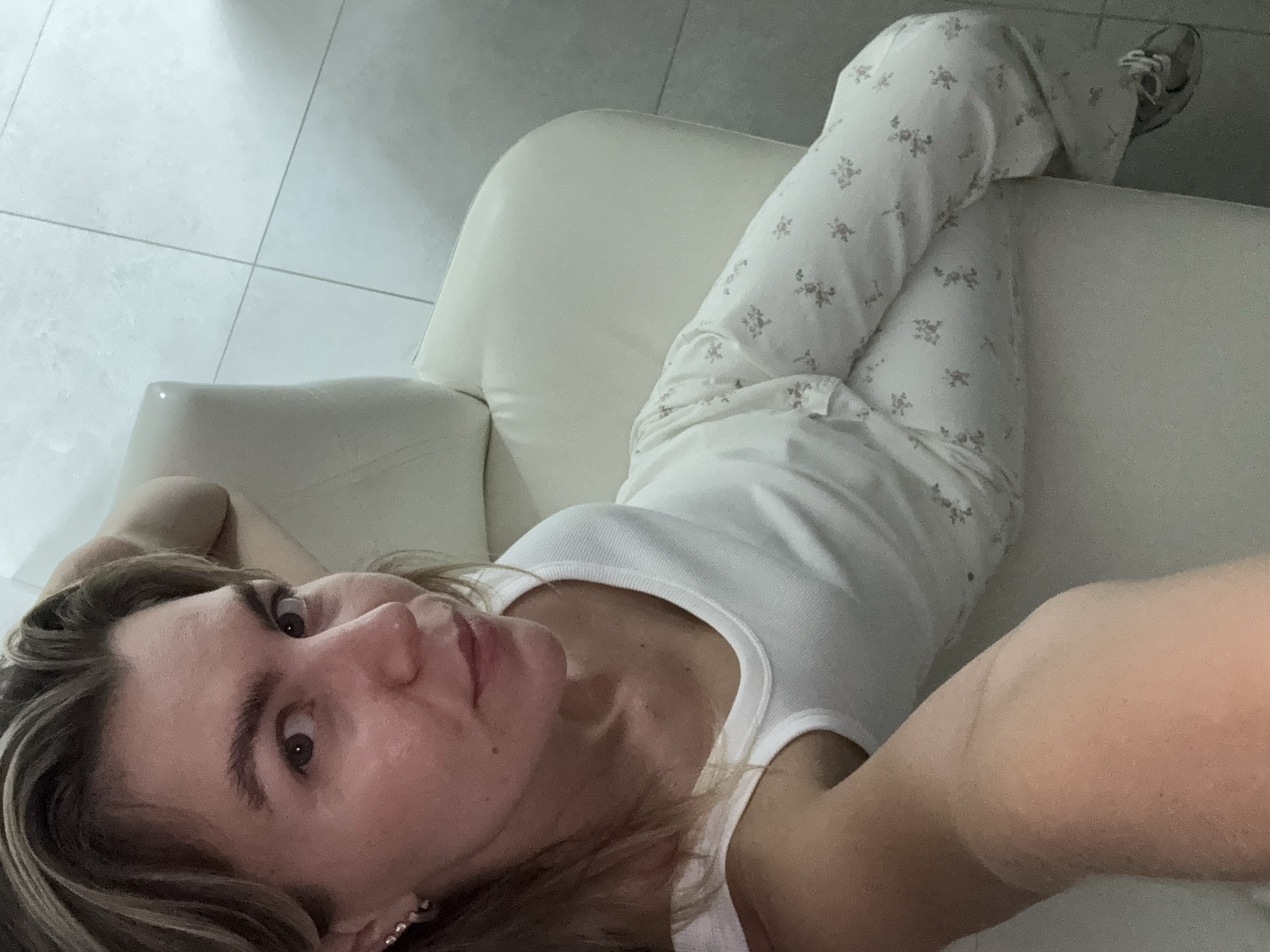 A woman is lying on a white couch with her head resting on her arm. She is wearing a white tank top and patterned pajama pants. The floor is tiled with a light color. The lighting is dim.