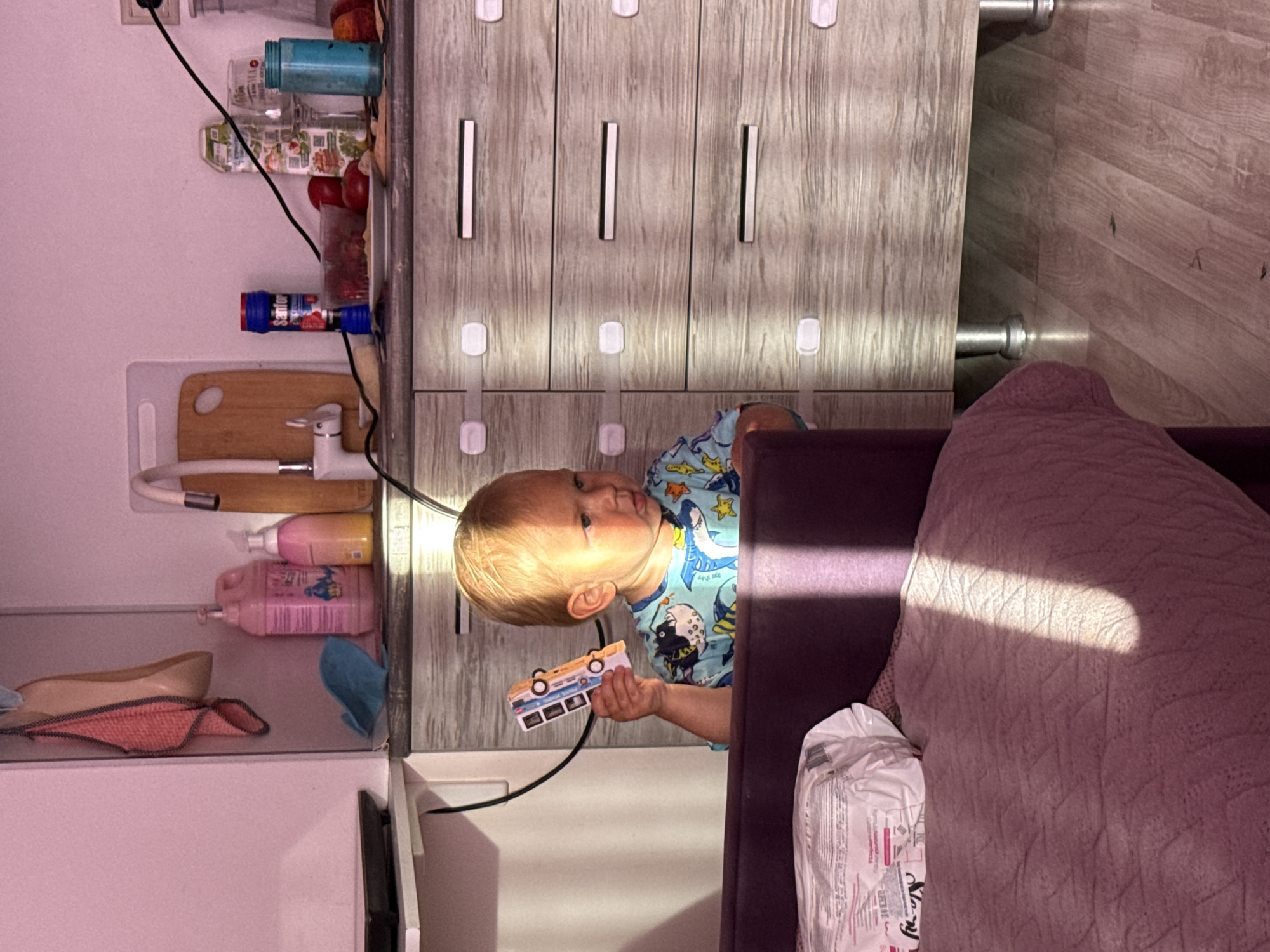 A young child with blonde hair, wearing a blue t-shirt with a shark and fish pattern, stands in front of a wooden dresser in a room that appears to be a kitchen or bathroom. The child is holding a toy bus in their right hand, looking to their right. The dresser has three drawers, each with a silver handle, and child safety locks on them. Sunlight casts a diagonal shadow across the scene from the left. To the left of the child, a purple upholstered furniture piece is visible, with a patterned blanket and a white bag on it. In the background, a white countertop features various items, including bottles of cleaning products, a cutting board with a faucet, a container of berries, apples, a carton of juice, and a blue jar. The wall behind the counter is pink. The flooring is light wood laminate. The image was taken indoors, likely in a residential setting. The location is not specified as Sochi, Russia.