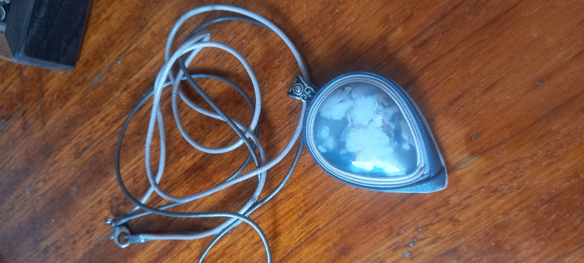 A pendant necklace with a teardrop-shaped stone is displayed on a wooden surface. The stone is pale blue and white, with a cloudy, almost floral pattern. The pendant is framed by layers of what appear to be blue and light grey material, possibly leather or suede. The necklace cord is made of two strands, one dark grey and one light grey, coiled loosely around the pendant. A small, ornate silver clasp is attached to the cord. The background is a polished wooden table with visible grain and some minor imperfections. There is no indication of the location being Izhevsk, Russia, or any other specific geographical area. The image is a close-up, overhead shot.
