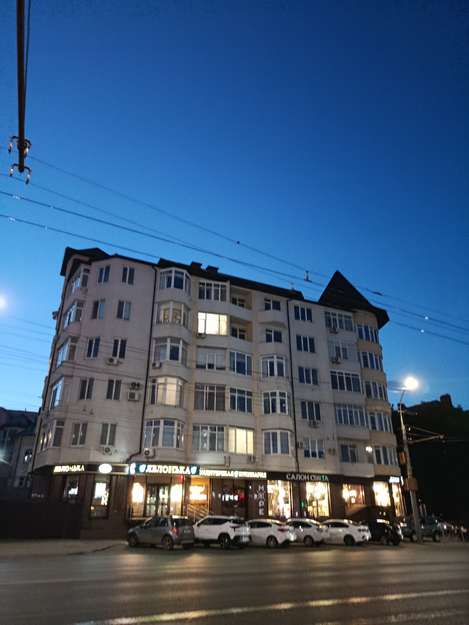 The image depicts a twilight scene in Saratov, Russia, focusing on a multi-story apartment building with illuminated storefronts on the ground floor. The building is a light-colored, modern structure with numerous windows and balconies. The storefronts are brightly lit, featuring signage that includes Russian text. Visible signs include "ЯБЛОНЬКА" (YABLONKA), "МОРИ", and "САЛОН СВЕТА" (SALON SVETA), suggesting a variety of businesses such as a shop, a cafe or restaurant, and a lighting store. Several cars are parked along the street in front of the building. Power lines are strung across the sky, and a street light is visible on the right side of the frame. The sky is a deep blue, indicating the transition from day to night. The overall impression is of a typical urban street scene in the evening.