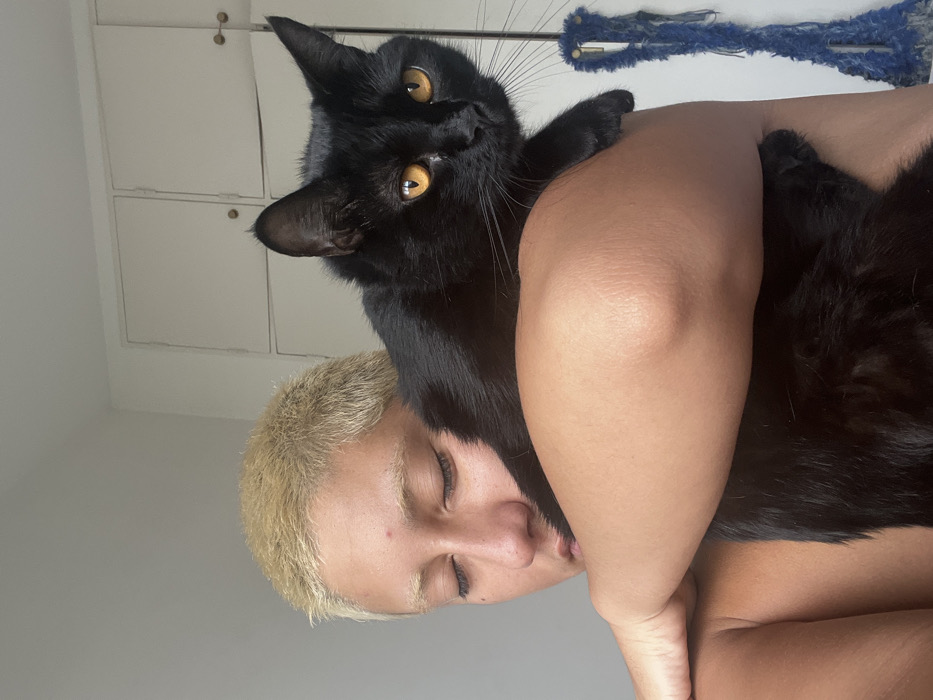 An indoor close-up photograph depicts an individual with a medium skin tone and short, bleached blonde hair, holding a black domestic cat. The individual's eyes are closed, and a small red blemish is present on their forehead. Their right arm is wrapped across their body, supporting the black cat on their right shoulder. The cat, with prominent amber-colored eyes, is facing towards the viewer and appears to be calmly resting. In the background, a light-colored wall is visible, featuring two light-colored cabinet doors with dark knobs in the upper right quadrant. A blue, textured, possibly frayed fabric item hangs vertically on the far right edge of the frame. The specific location within the city of Buenos Aires cannot be determined from the visual evidence in the image.