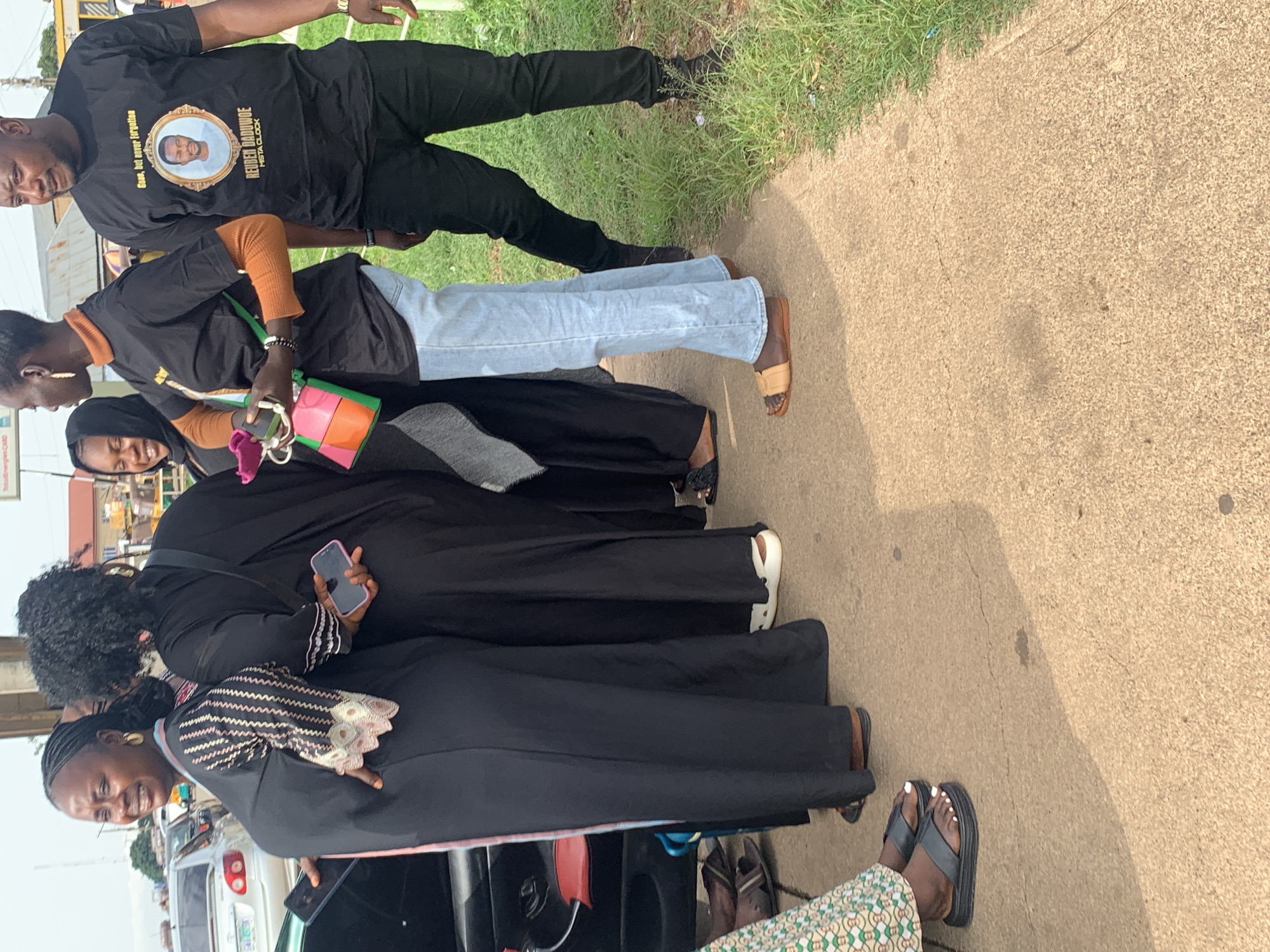 The image depicts a group of people standing on a sidewalk next to a road in Gura Top, Nigeria. On the left, a woman in a black abaya with patterned sleeves smiles while holding a dark blue phone. To her right, another woman in a black abaya holds a pink phone. A third woman in black, wearing a headscarf, smiles. Next to her, a woman in a black t-shirt with an orange turtleneck and light blue jeans holds a multicolored purse. The man to her right wears a black t-shirt with a portrait and text that reads "Gaas, but never forgotten. REUBEN DADUWOE. HISTA CLOCK." He also wears black pants. In the background, cars are visible on the road. The environment appears to be a commercial or residential area with a sidewalk and grass.