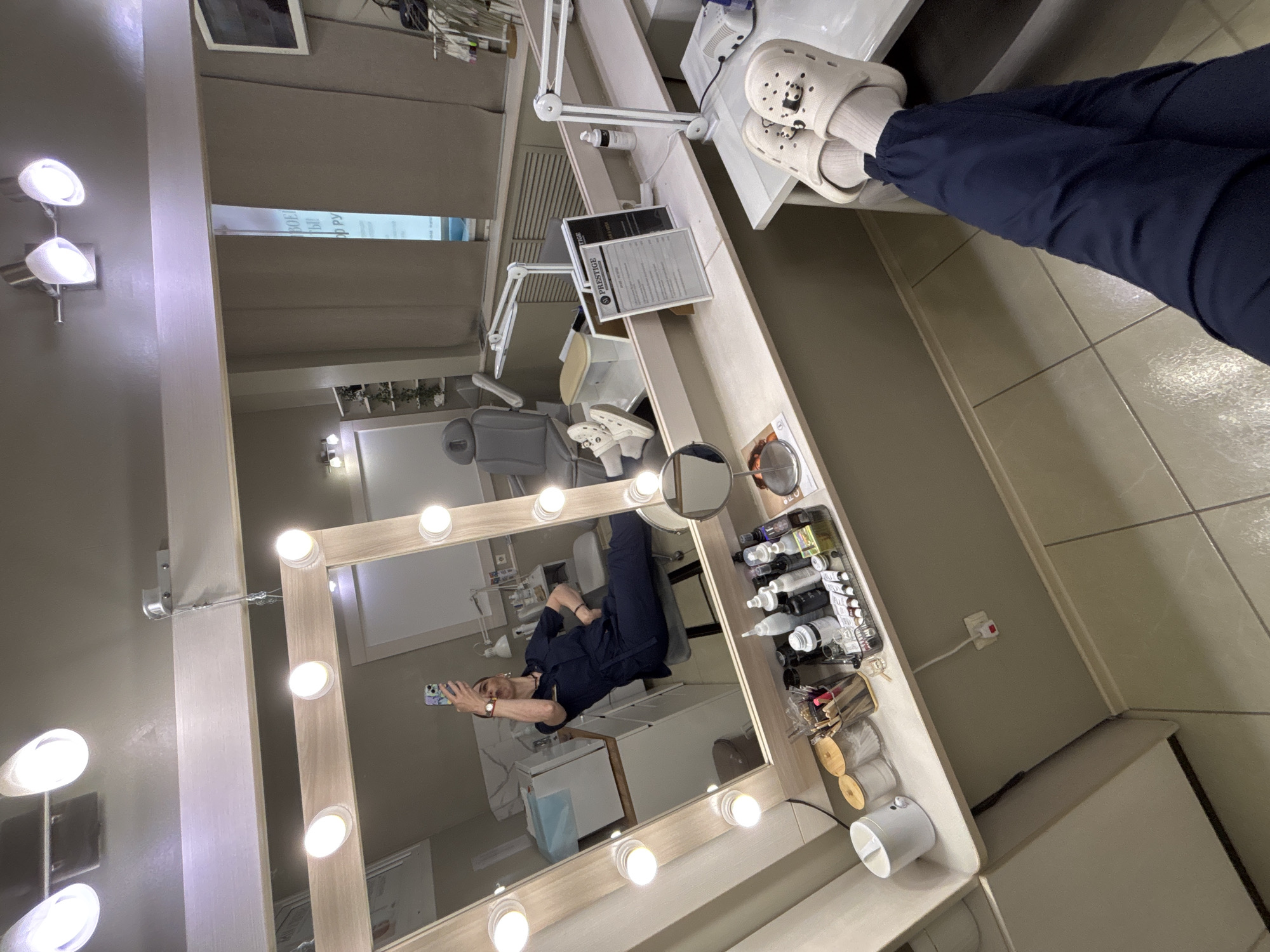 The image depicts the interior of a commercial or medical establishment in Vsevolozhsk, Russia, based on the signage in the background which includes Russian text. A mirror with illuminated light bulbs is prominent in the frame, reflecting a person holding a mobile phone in their right hand, presumably taking the photograph. The reflection also shows the interior of the establishment, including what appears to be a medical examination chair and various supplies. The scene includes shelves filled with bottles and containers.
The foreground shows a person's legs and feet wearing Crocs and socks. The angle and composition suggest the photo was taken in selfie style, capturing both the person's feet and the reflection of the room and the person themselves in the mirror. The environment seems sterile and clean, suggestive of a medical or cosmetic treatment center.