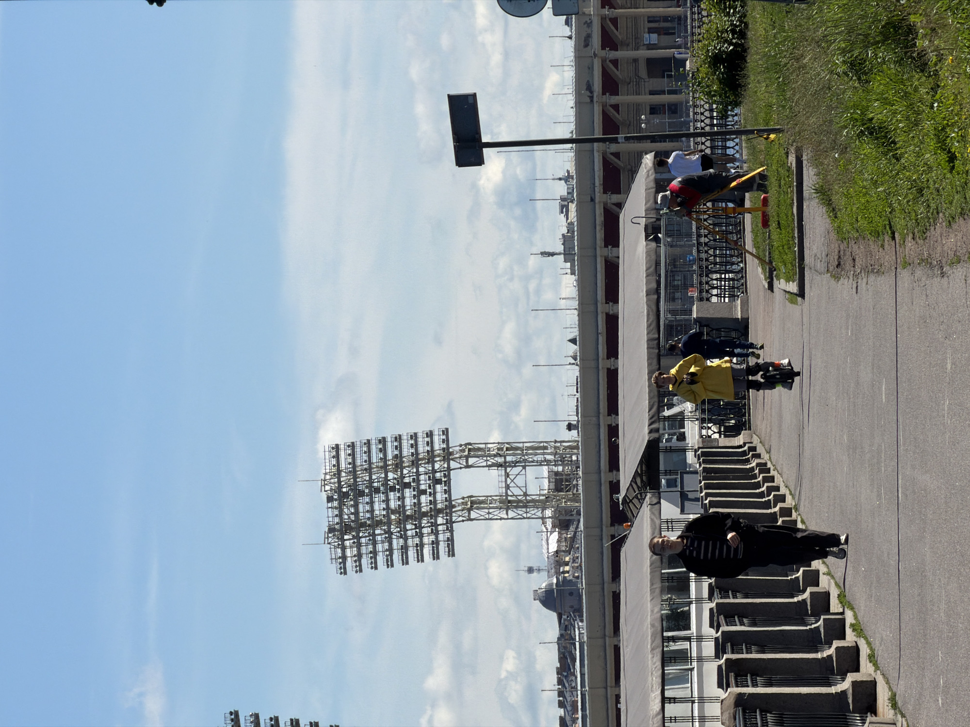 The image depicts a sunny day in Saint Petersburg, Russia. In the background, a tall stadium light structure is visible, along with the roof of a stadium and other buildings. In the foreground, a paved walkway runs alongside a river embankment.
On the walkway, several people are present. A man in dark clothing is walking towards the viewer, and another person is riding an electric unicycle. Further to the right, a person in a red and black uniform is operating surveying equipment on a grassy area next to the walkway. The embankment is lined with a decorative metal fence and stone posts. The sky is blue with scattered clouds.