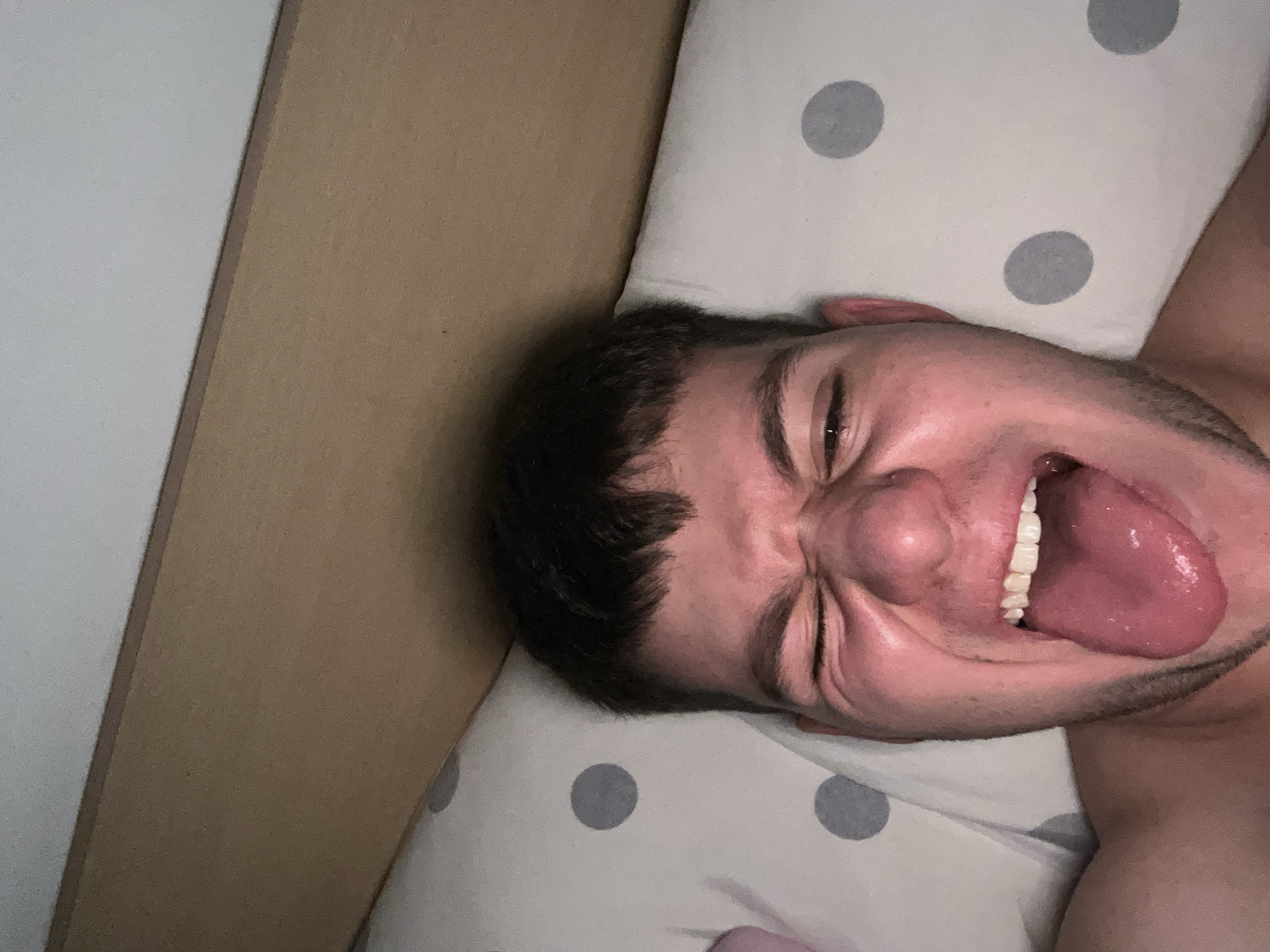 A man is lying on his back on a pillow with grey polka dots. He is sticking his tongue out and has his eyes closed. The background consists of a wall made of two different colored materials, one white and one beige. The location is not discernible from the image.