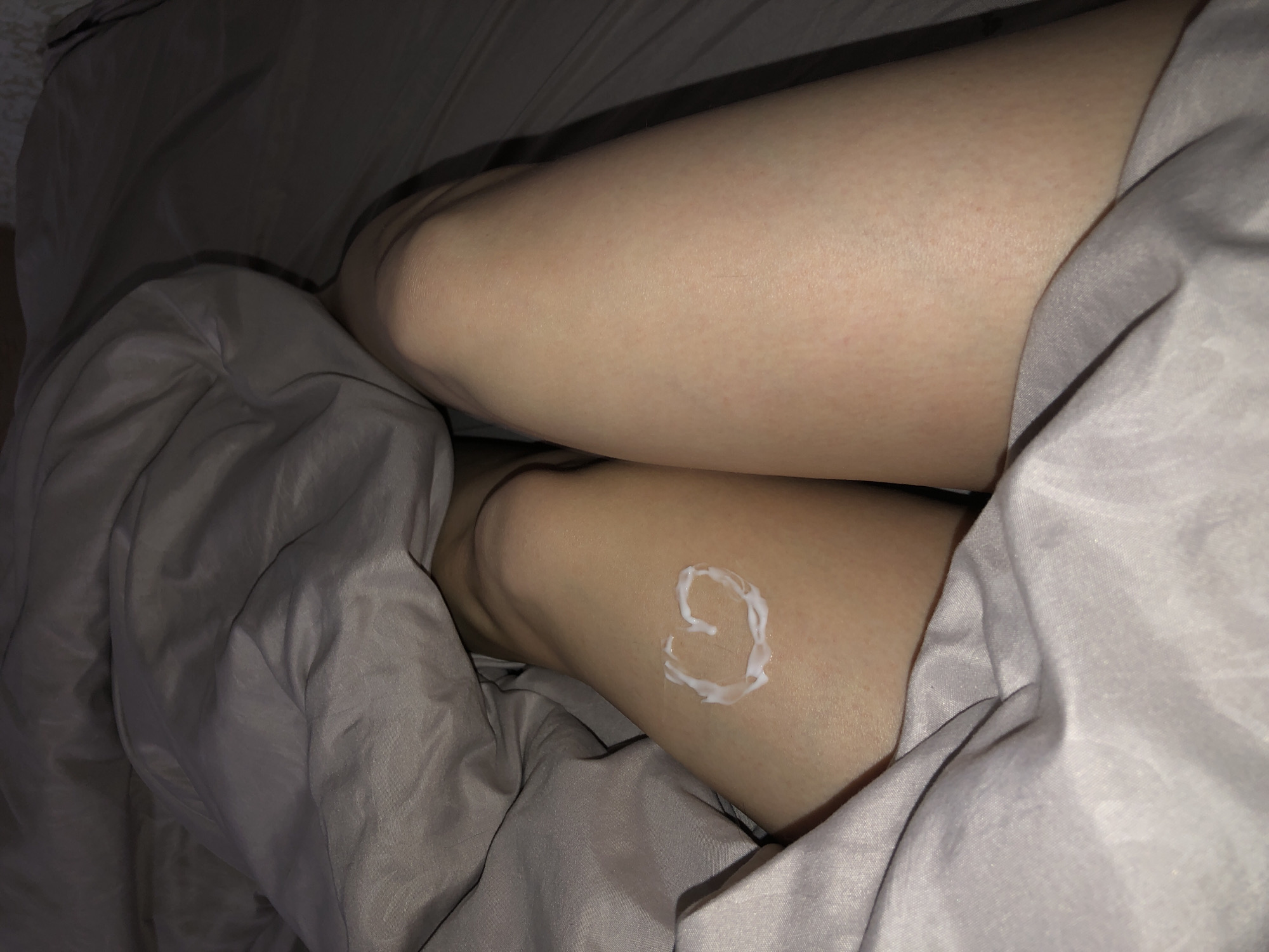 A person's legs are positioned in a bed covered with gray sheets. A heart shape made of a white substance, possibly lotion or cream, is on the lower right leg. The lighting is dim, casting shadows across the bedding. The scene appears to be indoors. The legs are pale skin toned. There is no evidence to suggest the image was taken in Noviy, Russia.