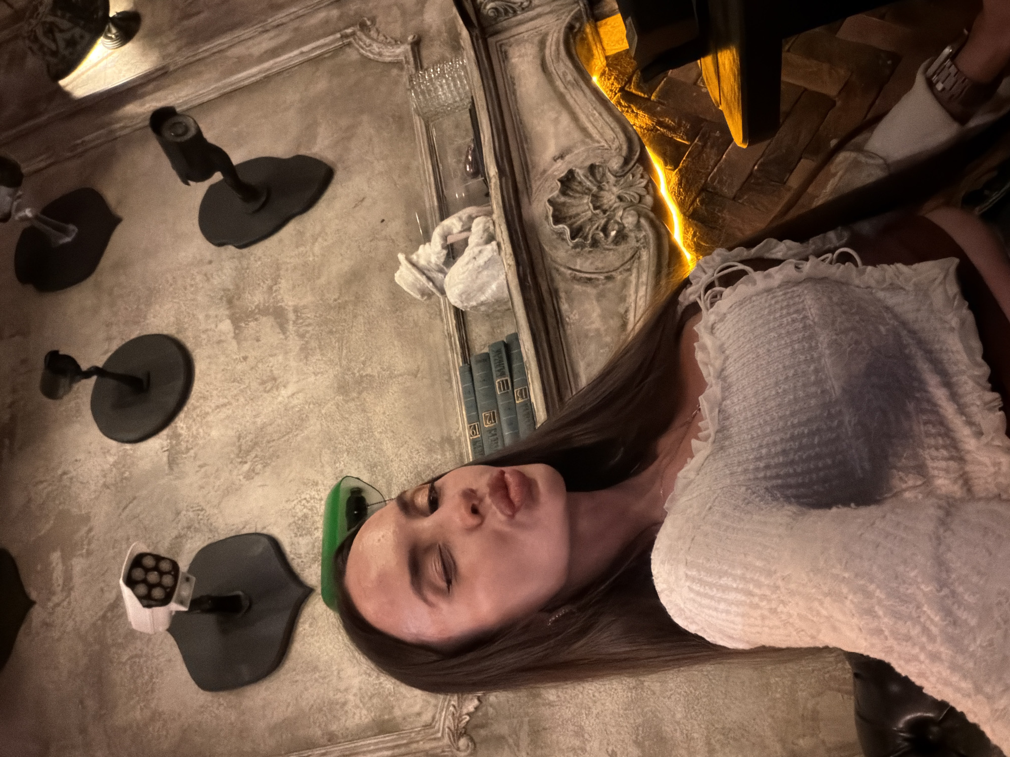 A young woman with long dark hair is taking a selfie in what appears to be a dimly lit, unconventional bar or restaurant in Saint Petersburg, Russia. She is wearing a white, textured top with ruffled details. Her lips are puckered in a kiss pose, and she is winking. The background features a textured, grey wall adorned with several vintage-style security cameras mounted on black plaques. To her right, a decorative fireplace mantle is visible, featuring a bust of a man in a hat, a stack of books, and a glass decanter. A strip of yellow light illuminates the area below the mantle, casting a warm glow on the brickwork. The overall ambiance suggests a themed establishment with an eclectic, possibly retro, aesthetic.
