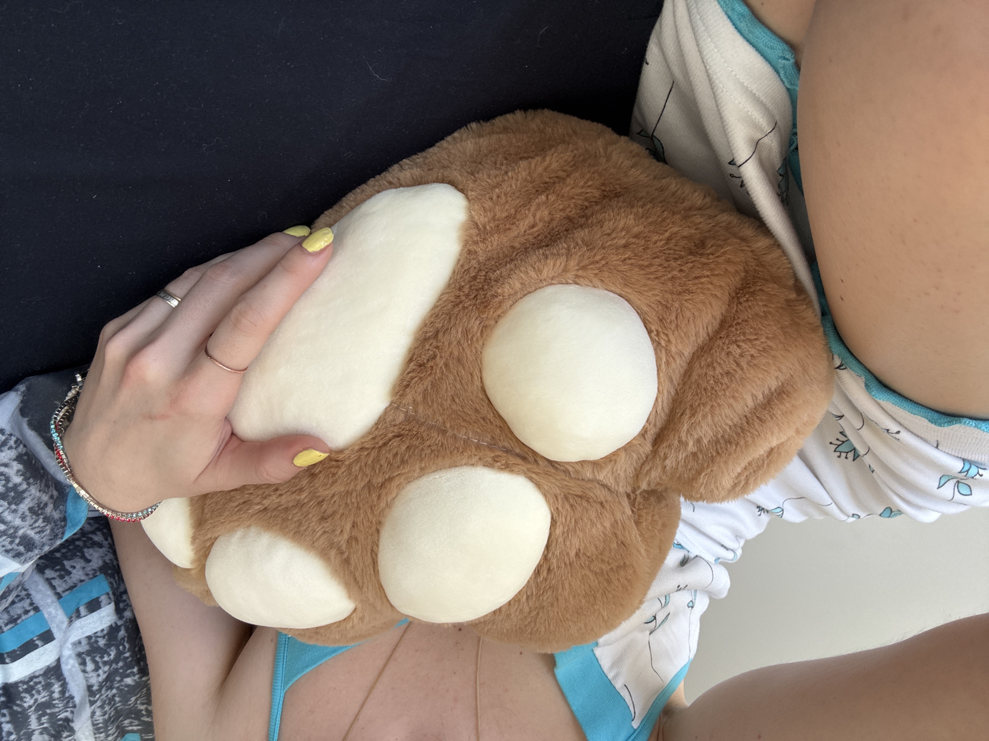 A close-up, high-angle shot shows a person holding a large, plush, brown bear paw. The paw has a white padded area representing the foot pad and toes. The person's hand, with yellow nail polish, rests on the paw. They are wearing a black top. Parts of the person's legs and clothing are visible. The clothing includes a white and blue top, white pajama-style bottoms with a blue pattern, and a blue bikini top. A bracelet adorns their wrist. The location cannot be determined from the visual information provided.