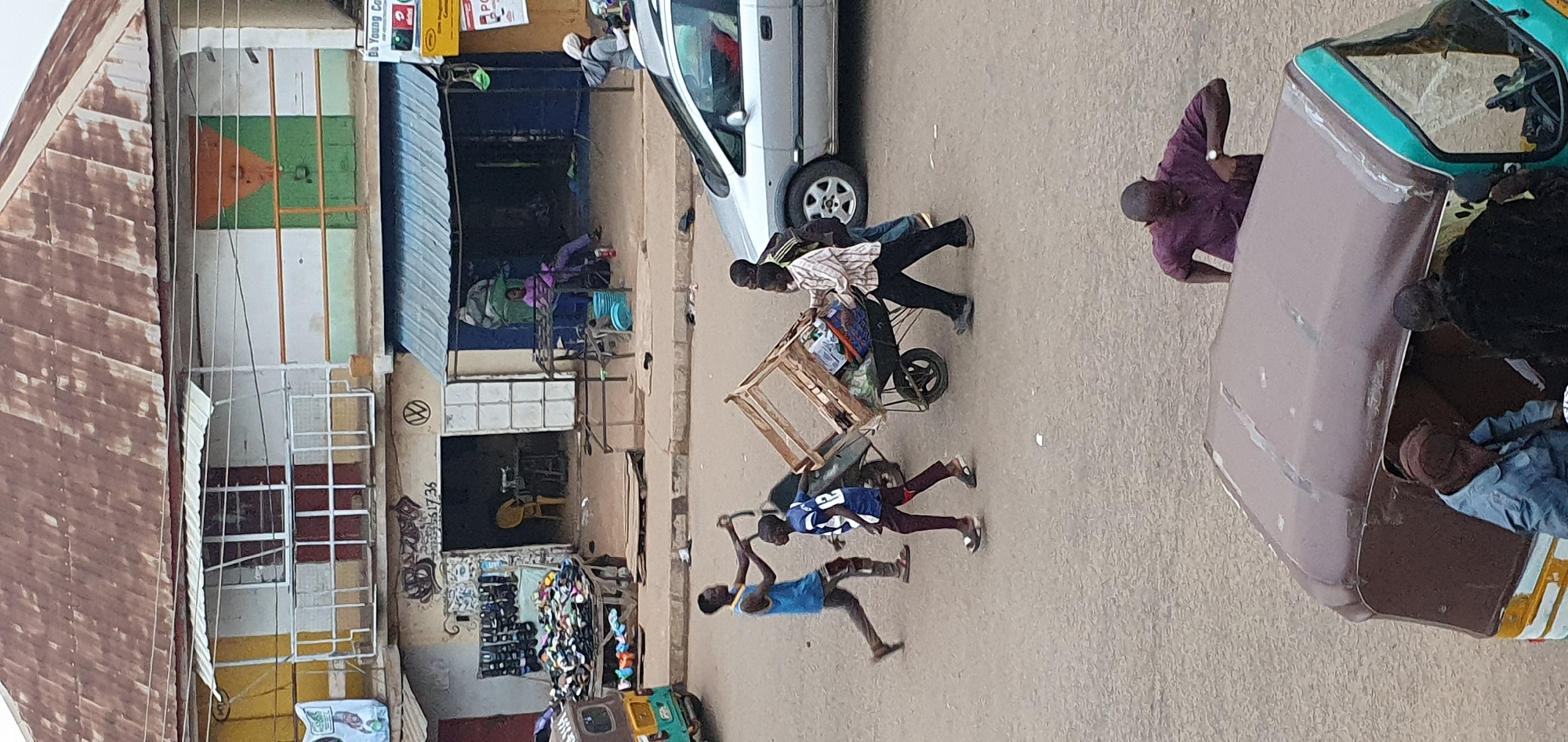This media file depicts a bustling street scene in Bukuru, Nigeria, likely during daylight hours under fair weather conditions. The focus is on the movement and activity of several individuals engaged in commerce and transportation.
The primary subjects are a group of people, including men and boys, in various states of motion. One man in a striped shirt and dark pants is pushing a wooden cart loaded with goods, accompanied by another person. Further down the street, two young boys are walking, one in a blue shirt and the other in a blue and white striped shirt, appearing to be involved in or observing the activity. In the background, other figures are visible, including one man in a purple shirt and another partially obscured by a motorized tricycle, suggesting a busy marketplace or thoroughfare.
Objects in the scene include a silver car, the wooden cart, and the motorized tricycle. The tricycle, with a light green and teal body, is parked or moving slowly, with people near it. The background features buildings with storefronts and corrugated metal roofs, indicative of a commercial area. One building has a sign with a Volkswagen logo. The ground is paved, suggesting a road or market square.
The overall activity appears to be the transport of goods and people, typical of a commercial street in a Nigerian town. The presence of multiple people and vehicles suggests a lively and active environment.
Visible text includes what appears to be a Volkswagen logo on a building, and some partially legible numbers and graffiti on another wall, possibly "51735". There are also posters on a building to the left, but the text is too small to read clearly. The scene conveys a sense of everyday commerce and community interaction.