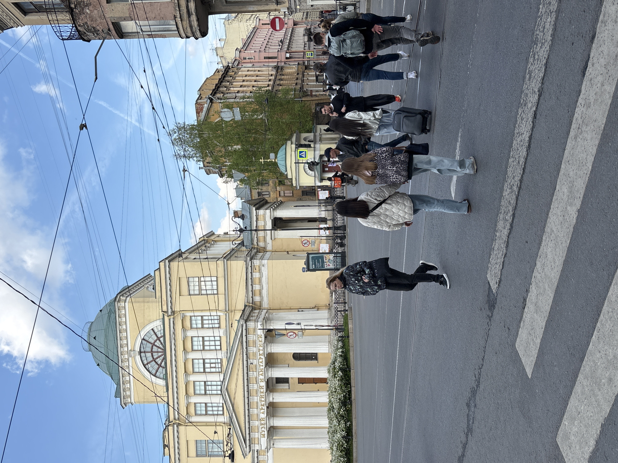 The image depicts a street scene in Saint Petersburg, Russia, showing a group of people crossing a pedestrian crosswalk. The architectural style of the buildings indicates a historic urban setting. The building on the left is light yellow, with a distinctive dome and classical columns. The building is identified as the Museum of Arctic and Antarctic. Overhead, there are numerous power lines crisscrossing the sky. The sky is light blue with a few clouds and jet contrails. On the right side of the street are buildings of varying heights and colors. At the end of the sidewalk, there is a "do not enter" traffic sign. The group of people is diverse in terms of age, dress, and ethnicity. Some individuals are wearing backpacks or pulling suitcases, suggesting they may be tourists. They are actively crossing the street, moving from right to left in the frame.