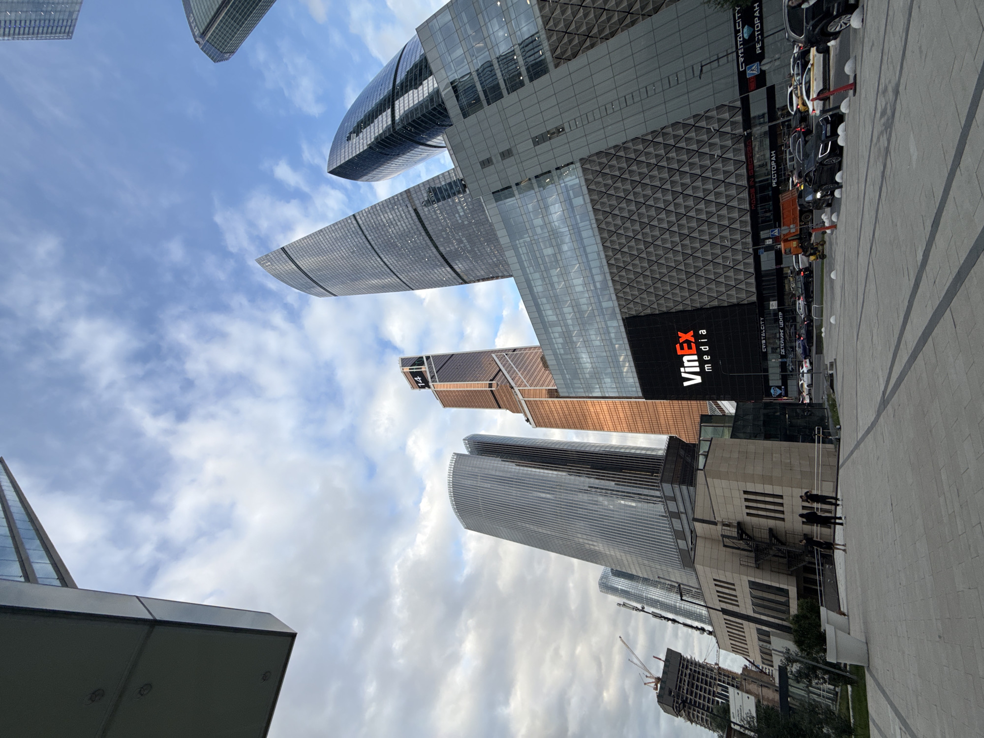The image depicts a modern urban scene with several skyscrapers piercing a cloudy sky. The architecture is characterized by sleek glass facades and geometric patterns, suggesting a contemporary business district. In the foreground, a paved walkway leads towards the buildings, with a few people visible, seemingly observing the structures or conversing. One of the buildings features a prominent orange and black sign with the text "VinEx media". Other text, in Russian, is visible on signs including "РЕСТОРАН" and "ДЕТСКИЙ ЦЕНТР". Cars are parked along the street or in a queue near the buildings. Construction cranes are also visible, indicating ongoing development in the area. The overall impression is one of a bustling, modern city environment, likely during daytime given the lighting. The presence of multiple large office buildings suggests a commercial hub.