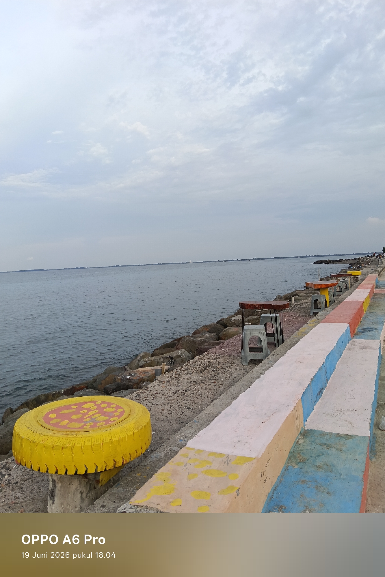The image depicts a waterfront scene in Lhokseumawe, Indonesia, on June 19, 2026, at 18:04. A calm sea stretches to the horizon under a cloudy sky. The foreground features a textured concrete walkway with colorful painted sections in blue, orange, and yellow. Several concrete benches, also painted, line the walkway. Further along, there are small tables and chairs, suggesting a seating area for visitors. A breakwater made of large rocks separates the walkway from the sea. To the left, a yellow, circular object, possibly a repurposed tire painted yellow with red and yellow patterns, serves as a unique seating element or table. No people are clearly visible in the immediate foreground or midground. The overall atmosphere is serene and somewhat utilitarian, designed for public use by the water.