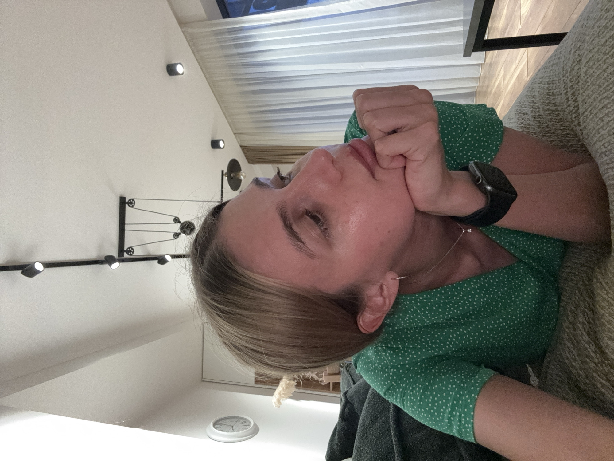 The image shows a woman lying down on her side, propping her head up with her right hand, which is clenched into a fist and covers her mouth and chin. Her eyes appear to be closed or looking upwards. She has light brown hair and is wearing a green top or dress with white polka dots. A silver necklace with a small pendant is visible, and a dark smartwatch with a silver-colored casing is on her left wrist.
The setting appears to be an indoor space with white walls and ceiling. Modern black track lighting fixtures are installed on the ceiling and a wall. A decorative wall-mounted object resembling a system of pulleys or gears is visible behind her. In the upper right corner, a window with horizontal blinds is partially visible. A patterned, light-colored fabric (possibly a blanket or sofa upholstery) is next to her, and a darker grey fabric is under her left arm. A small, round, white object, possibly a clock face, rests on a surface in the lower left.
The activity suggests rest, contemplation, or perhaps boredom. The indoor lighting is on, implying it could be evening or a room that requires artificial light. No visible text is present.